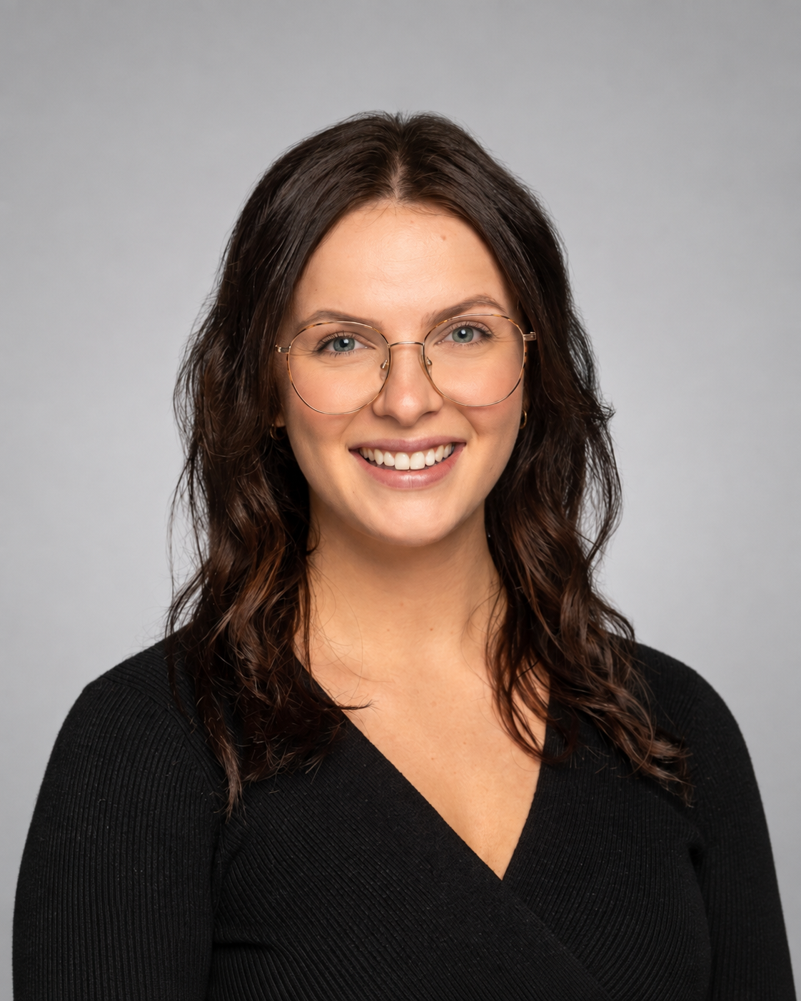 A woman with wavy brown hair wearing glasses and a black top, smiling against a plain gray background.