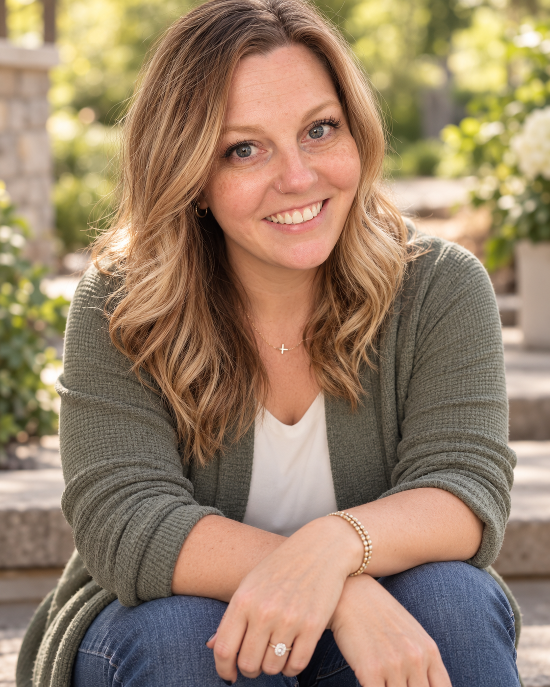 A woman with wavy blonde hair and blue eyes smiling outdoors, wearing a green cardigan, white top, bracelet, and ring, with greenery and flowers in the background.