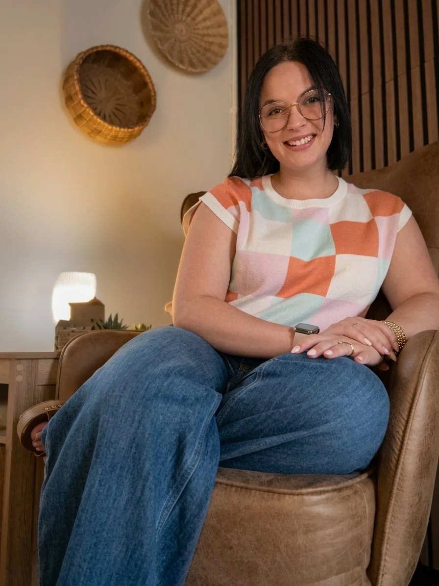 A woman sitting on a brown armchair, smiling, wearing glasses, a colorful patchwork top, and jeans, with wall decorations behind her.