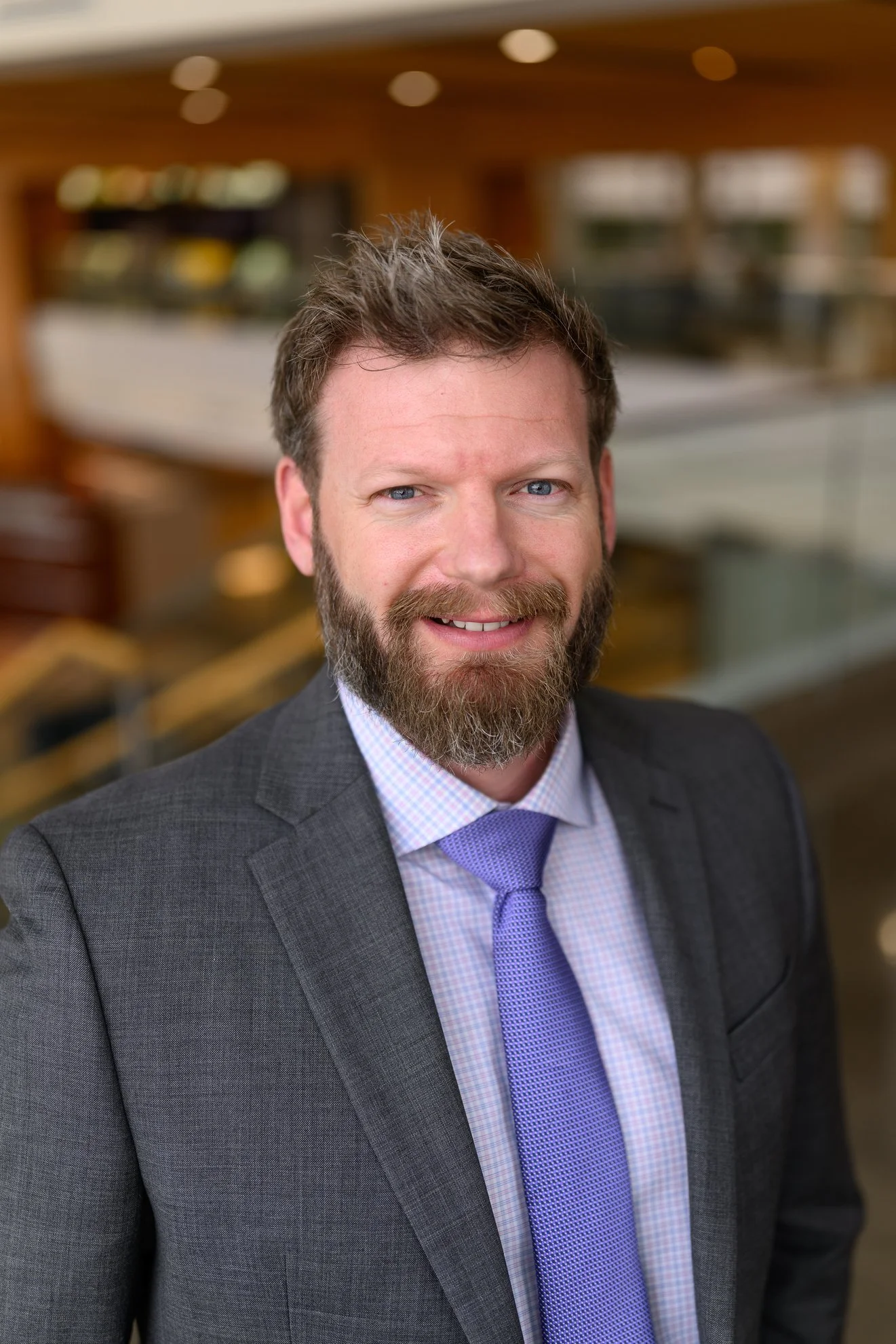 A man in a dark gray suit with a purple tie is smiling in an indoor setting.