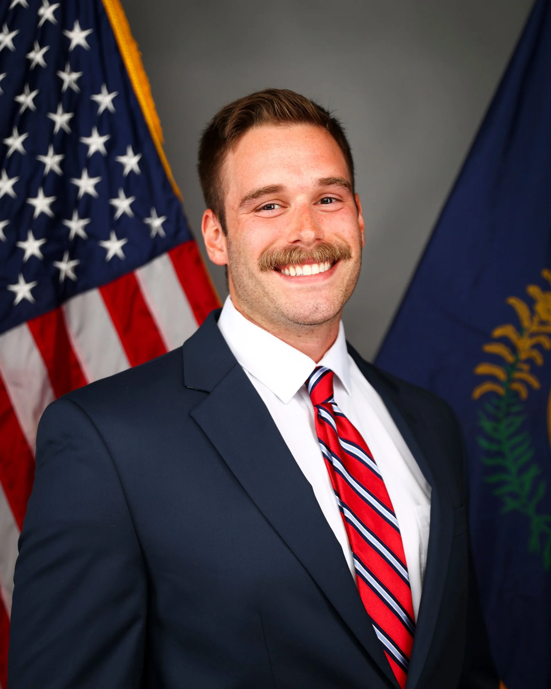 A man with short brown hair, a mustache, wearing a navy suit, white shirt, and a red, white, and blue striped tie, smiling in front of an American flag and another flag with a seal.