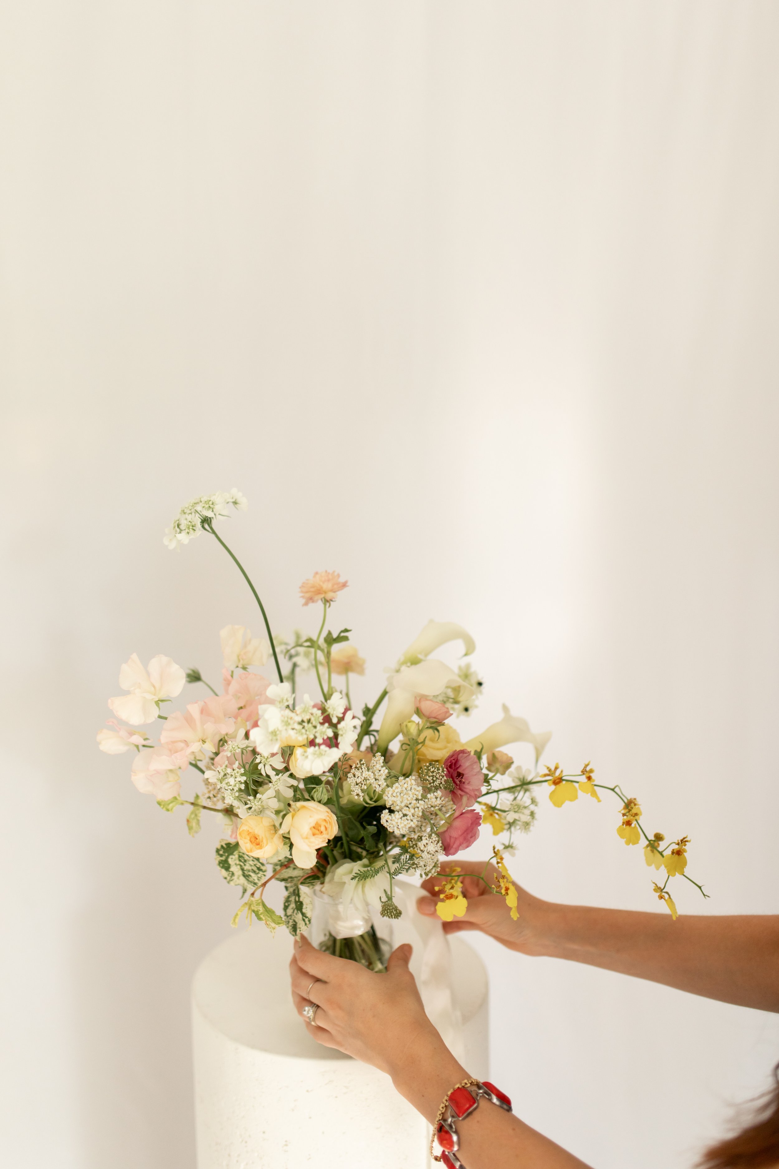 Person arranging a mixed flower bouquet in a white vase on a pedestal against a plain white background.