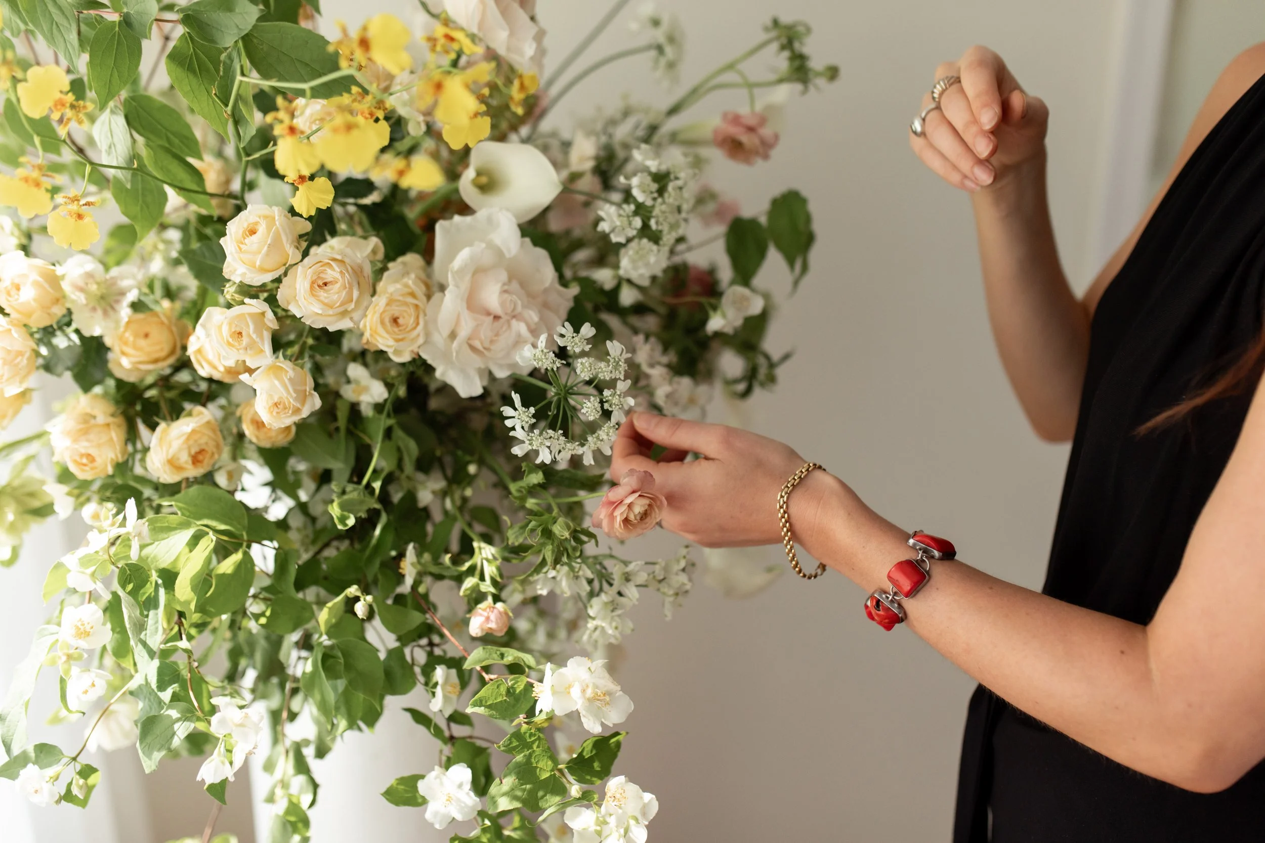 Person arranging a large bouquet of white and yellow flowers with green leaves, wearing rings, bracelets, and a black sleeveless top.