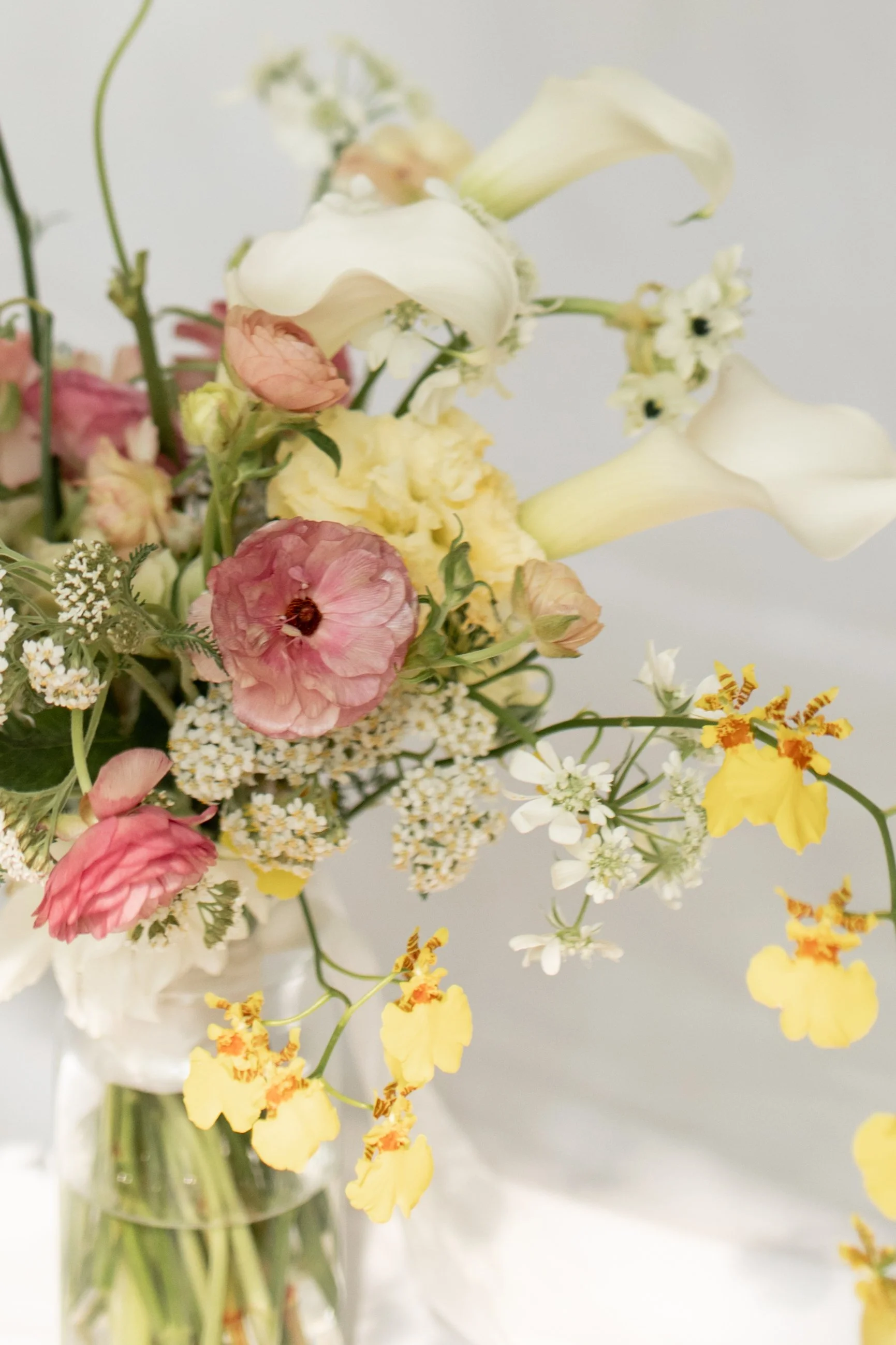 A bouquet of mixed flowers including white calla lilies, pink ranunculus, yellow orchids, and small white blossoms in a glass vase.