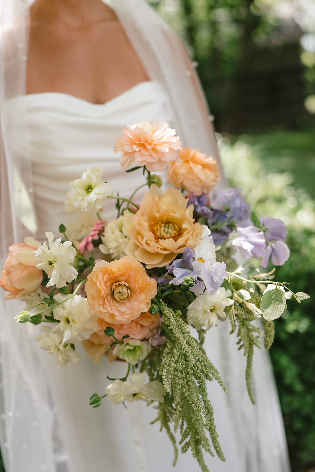 Close-up of a bride holding a colorful bouquet of roses, peonies, and greenery, outdoors with blurred greenery background.
