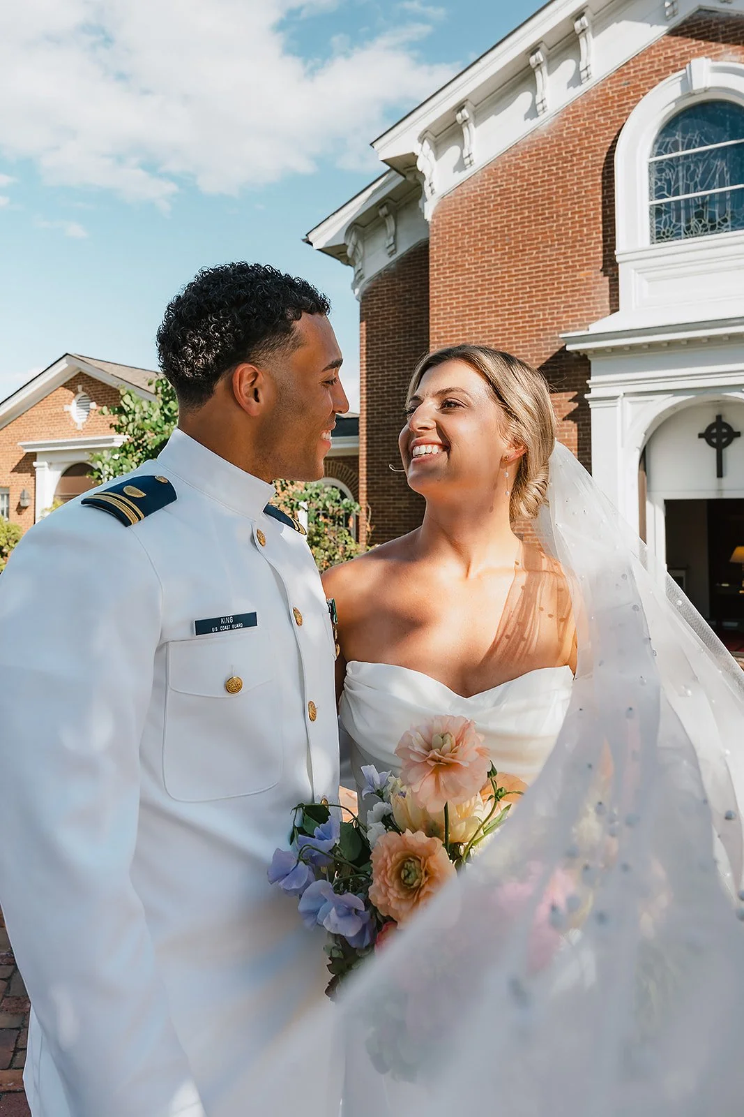 A bride and groom stand close together, smiling at each other outside a brick building with white decorative trim, under a blue sky with clouds. The groom is wearing a white military uniform, and the bride is in a white wedding gown with a veil and h