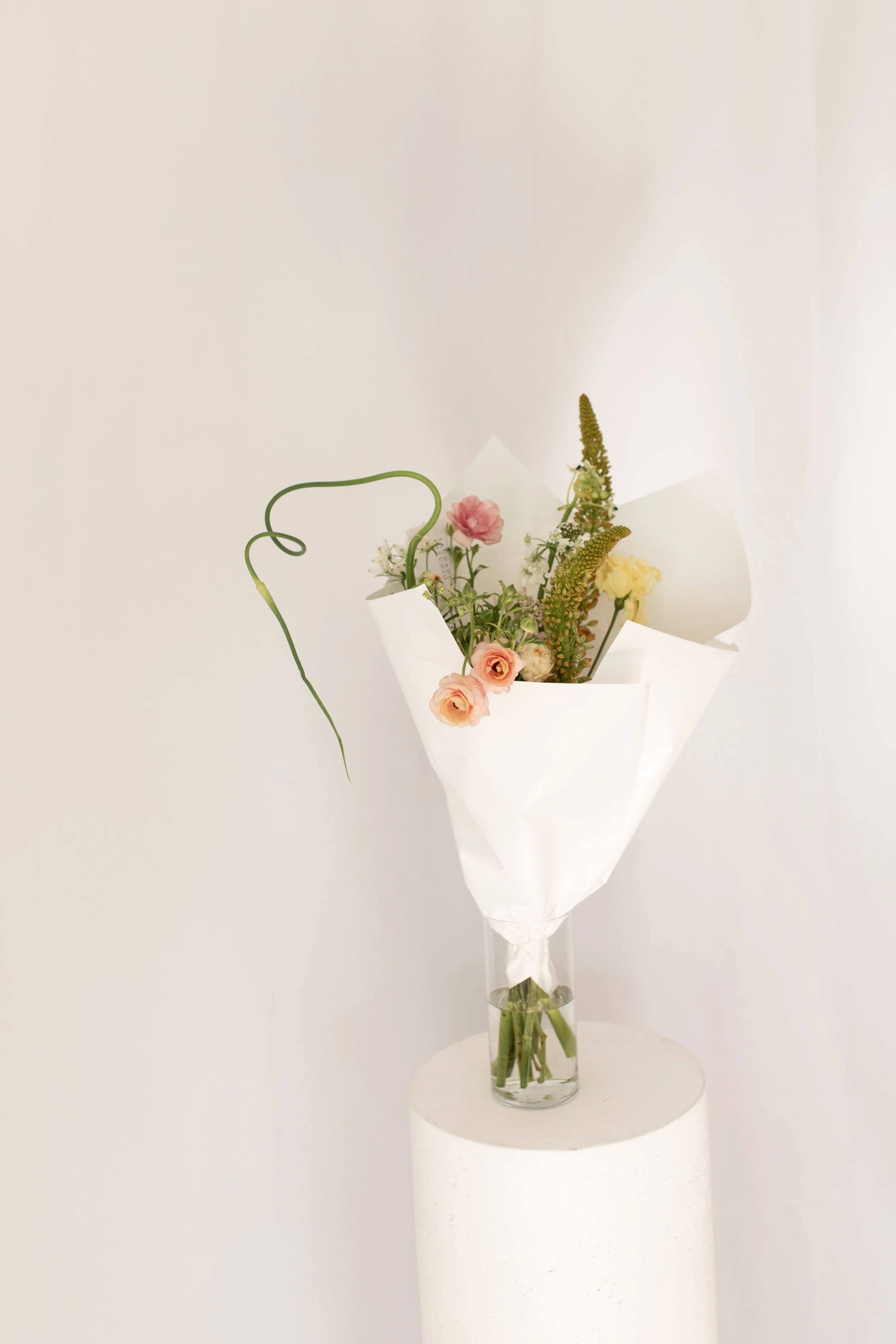 A bouquet of mixed flowers in a clear glass vase on a white pedestal against a plain light-colored background.