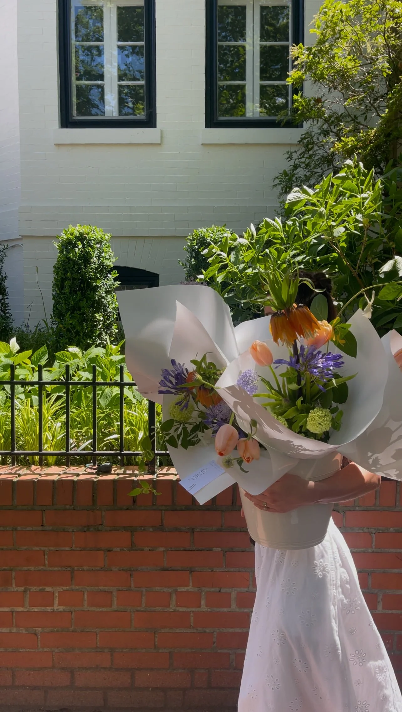 A person holding a large bouquet of flowers outdoors, with a brick wall, black metal fence, and a white building with green window frames in the background.