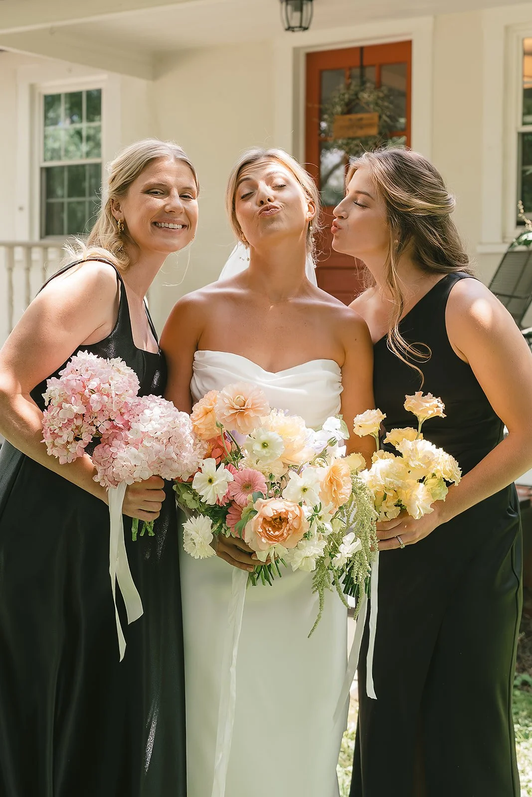 A bride in a white strapless dress holding a bouquet of light pink, white, and peach flowers, flanked by two women in black dresses each holding a smaller bouquet, standing on a porch outside a house, making kissing faces.