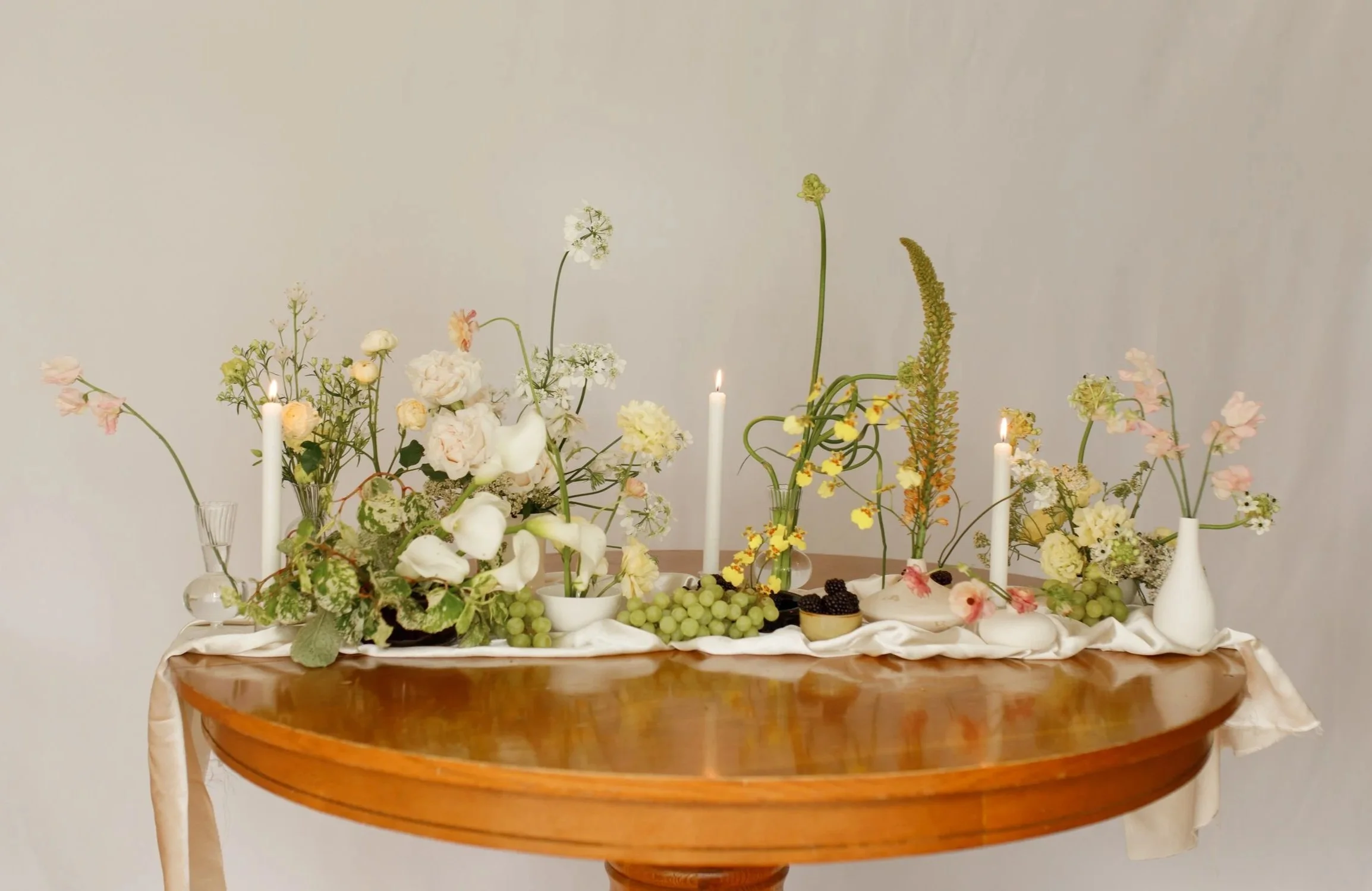Floral arrangement with white, pink, and yellow flowers, candles, and green grapes on a wooden table with a white cloth.