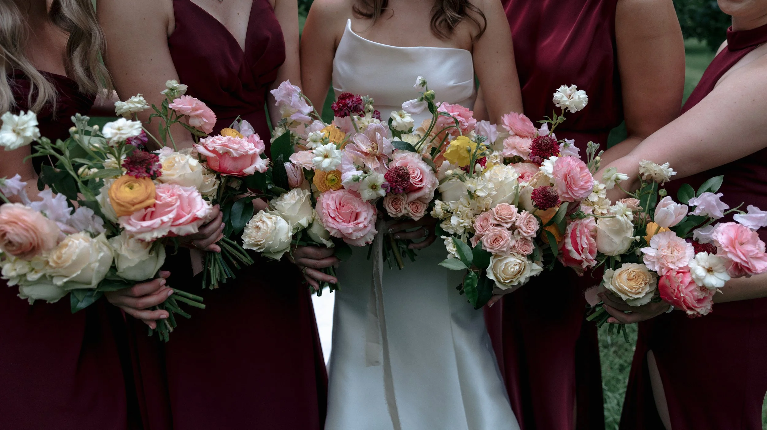 Bride in a white dress and bridesmaids in maroon dresses holding large bouquets of pink, white, and yellow flowers.