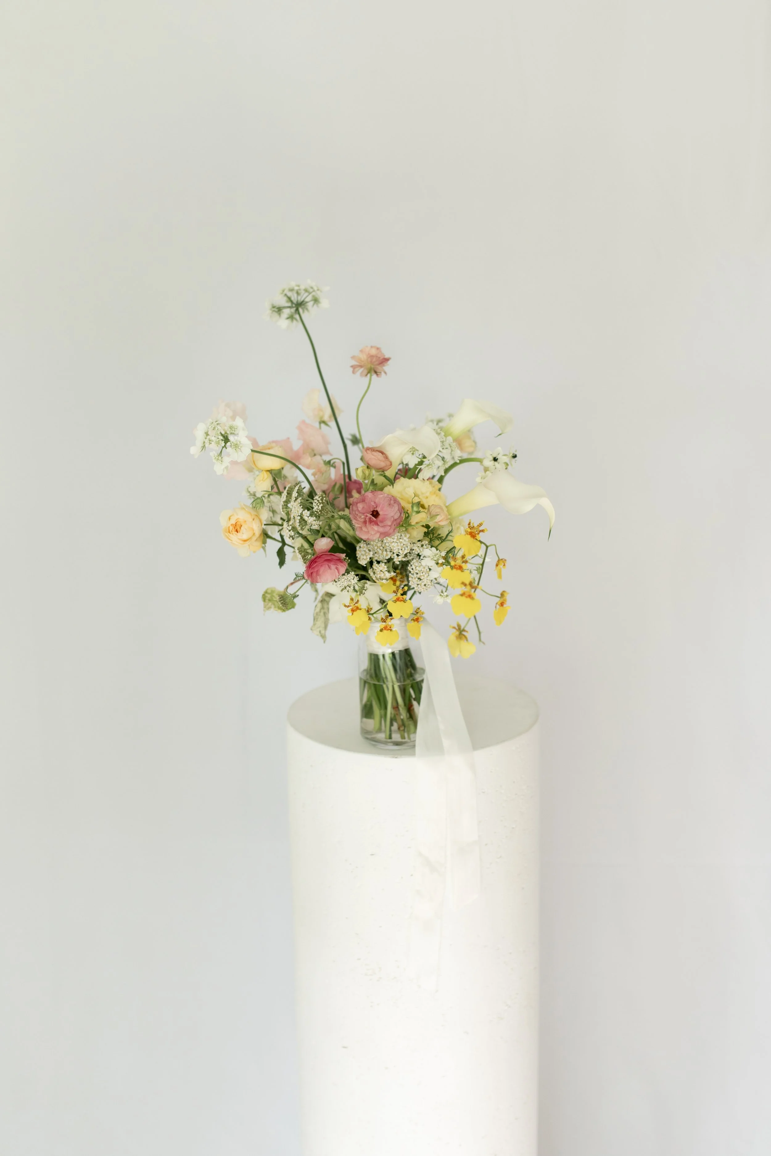 A bouquet of mixed flowers in a glass vase on a white pedestal, featuring calla lilies, roses, and other blooms against a plain white background.