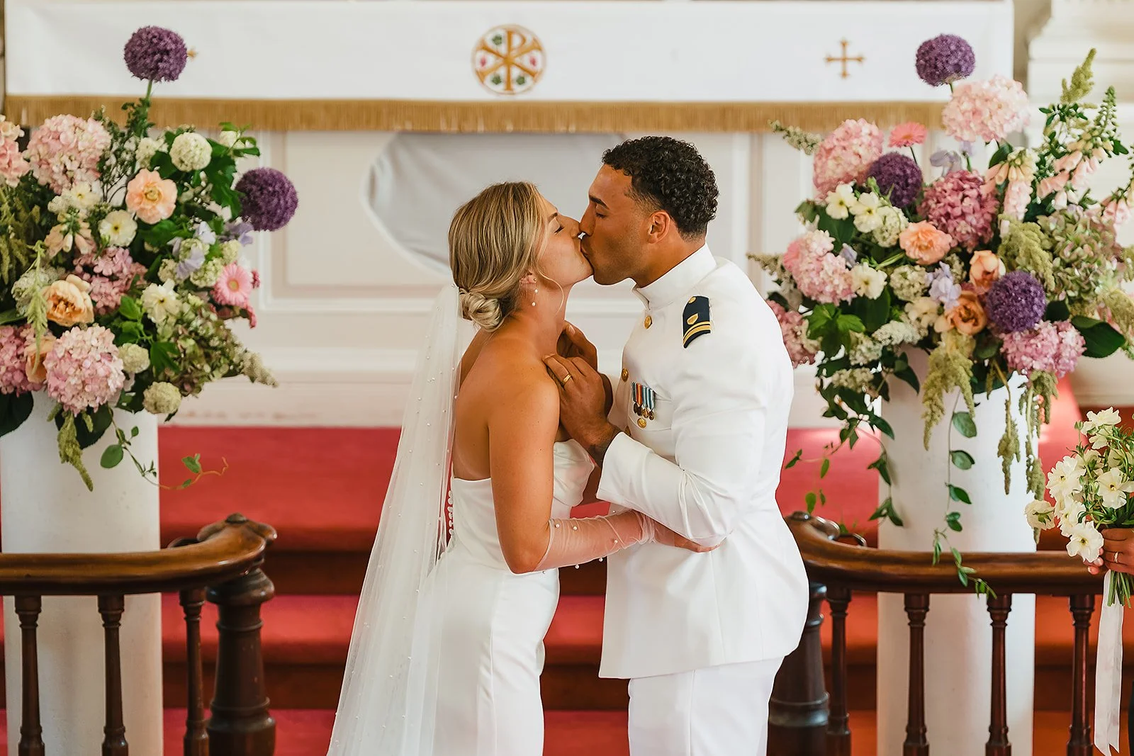 A bride and groom share a kiss during their wedding ceremony in a church decorated with large pink and purple floral arrangements.