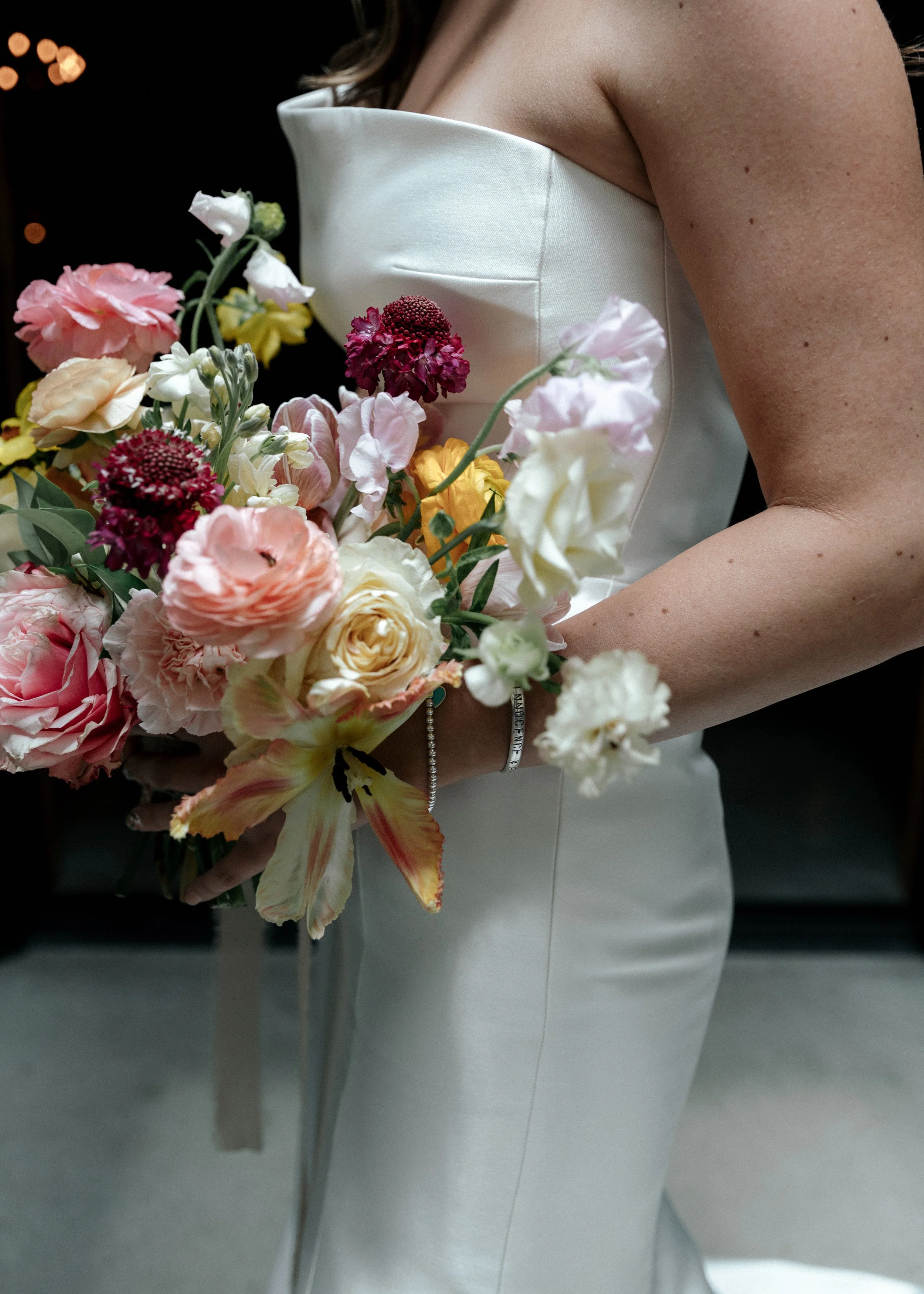 Close-up of a bride holding a bouquet of pink, white, yellow, and purple flowers, wearing a white strapless wedding dress and bracelet.
