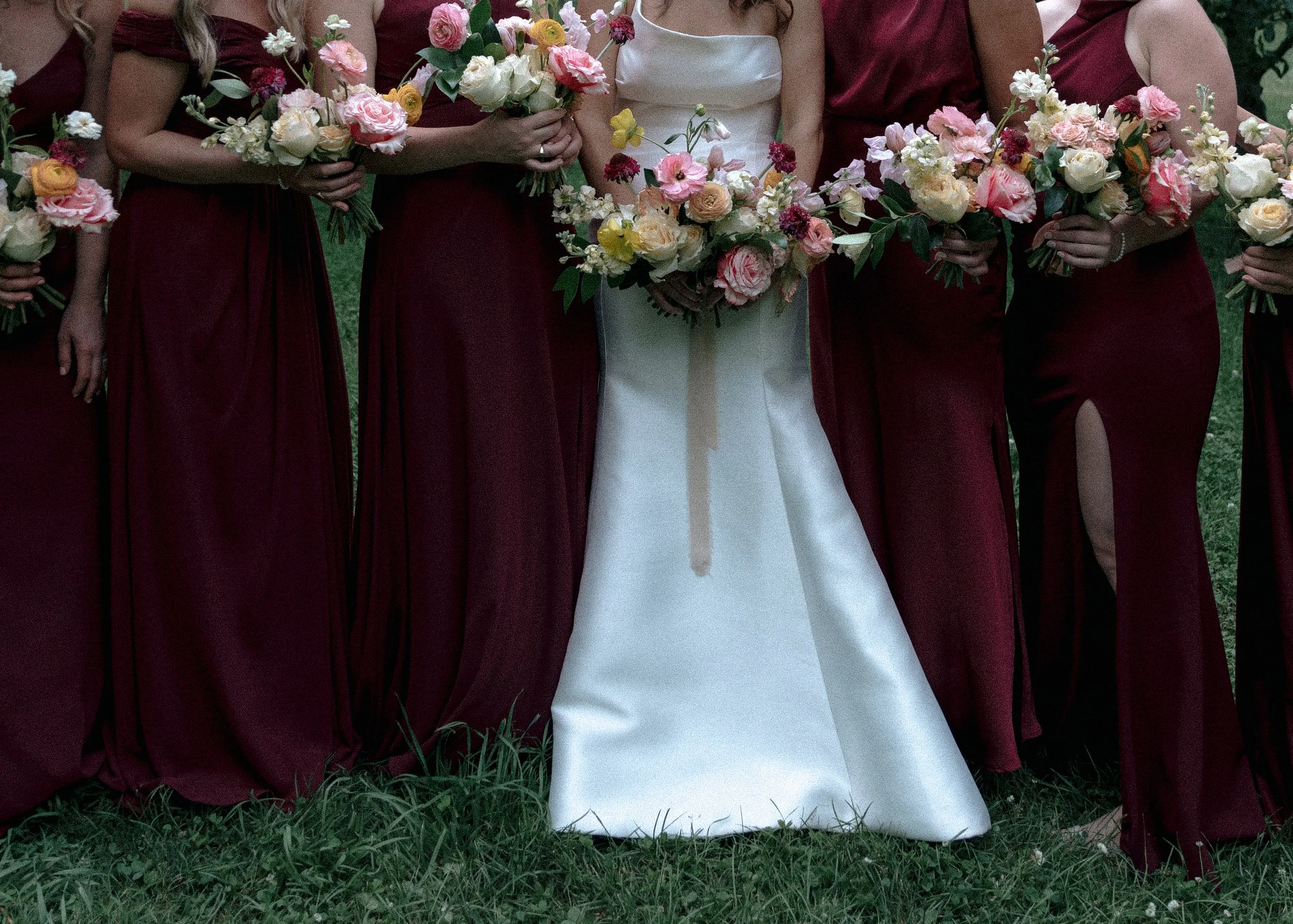 A group of women in burgundy dresses holding bouquets of pink, white, and yellow flowers, with one woman in a white wedding gown, standing on grass.