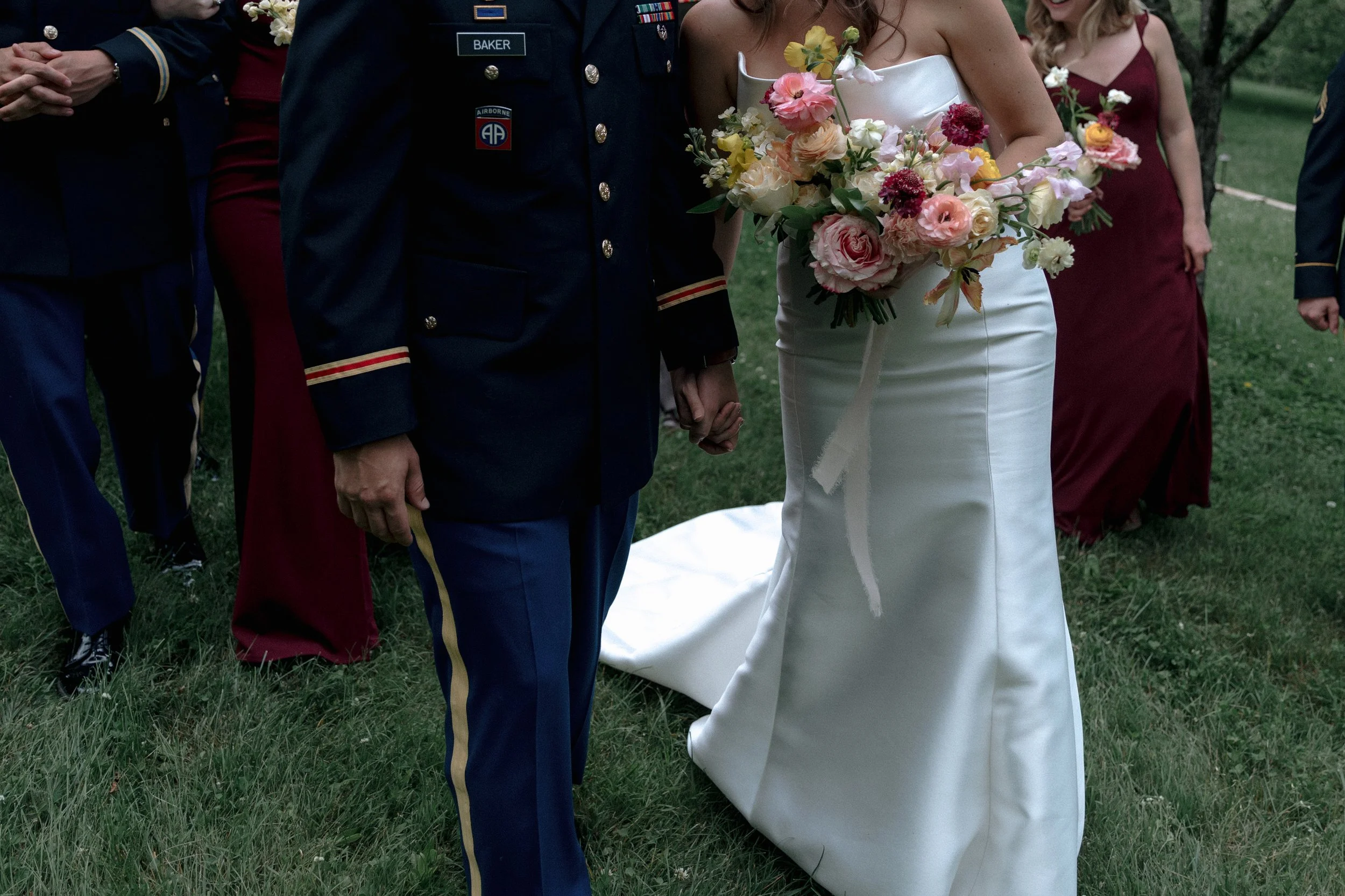 Bride and groom holding hands at an outdoor wedding ceremony, with bridesmaids and groomsmen in the background on a grassy area.