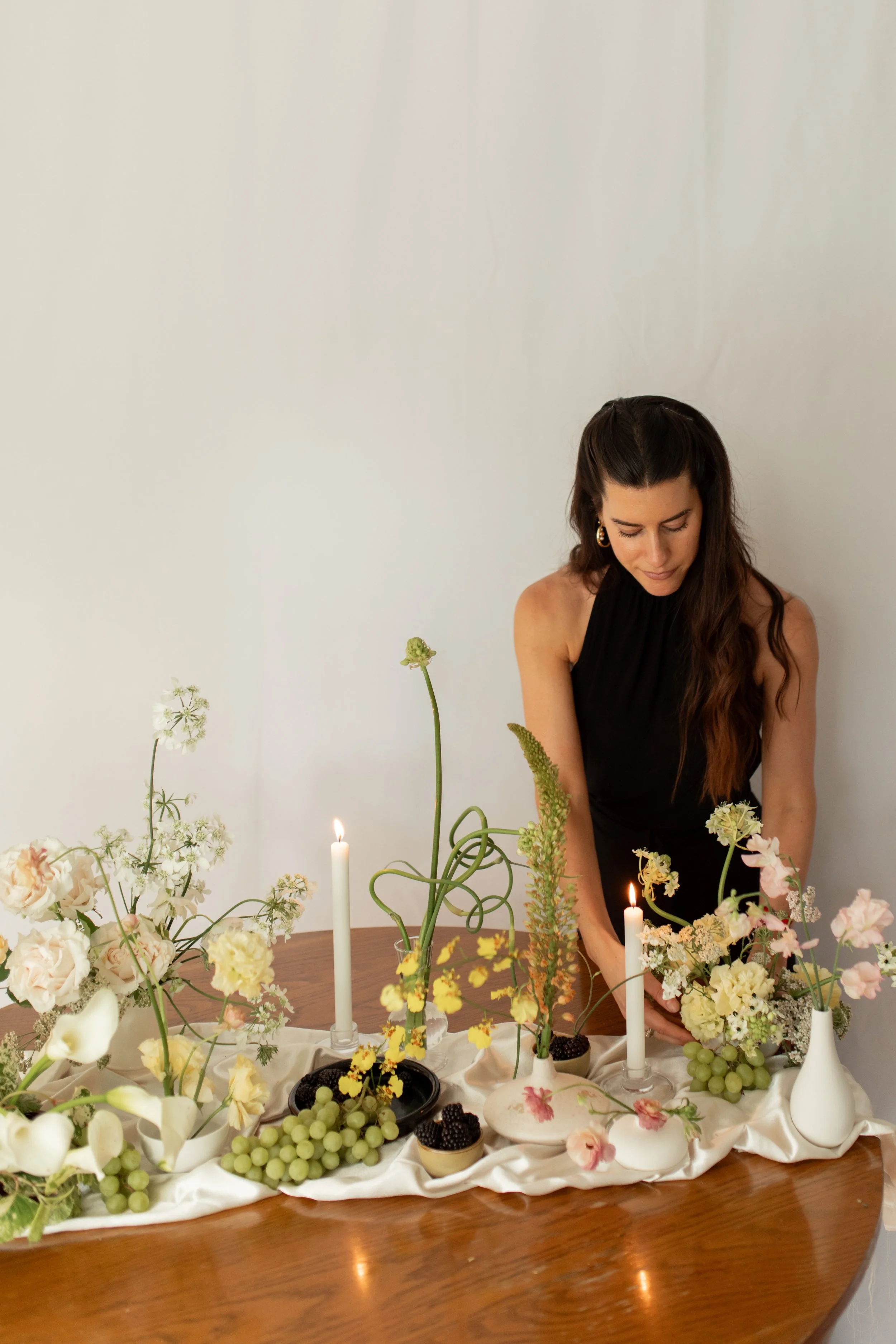 Woman arranging flowers on a table decorated with flowers, grapes, candles, and decorative bowls.