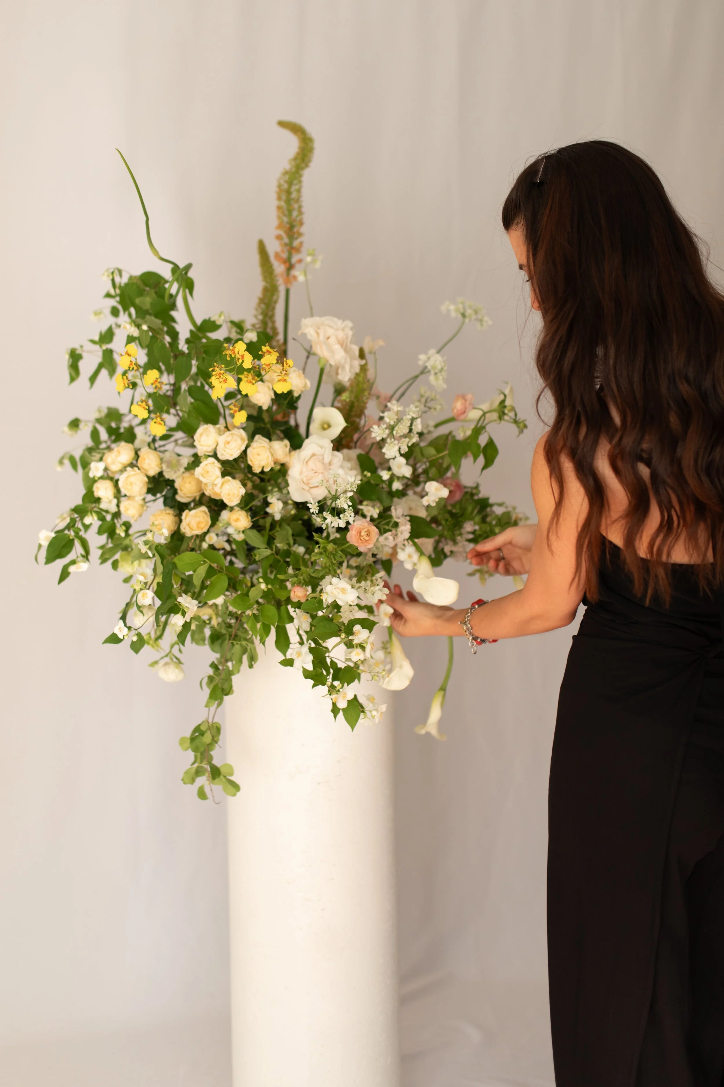 A woman in black dress arranging a large bouquet of white, yellow, and pink flowers in a white vase.