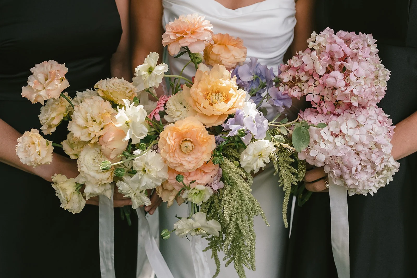 Two people holding bouquets of pastel-colored flowers including roses, hydrangeas, and other blossoms, with ribbons, during a wedding ceremony.