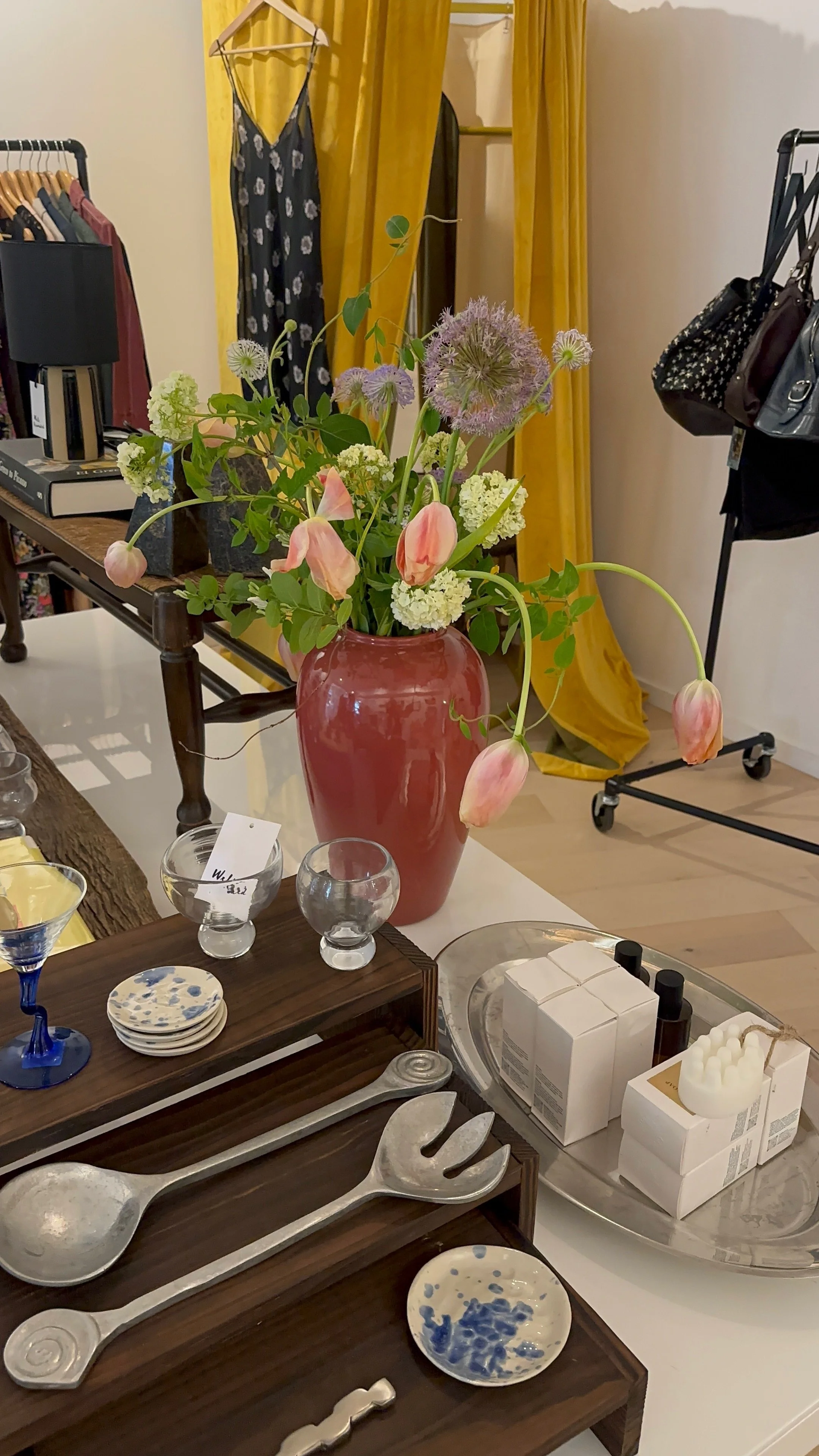 A store display with a pink ceramic vase filled with pink tulips, white hydrangeas, and purple alliums. Surrounding objects include plates, glassware, decorative items, and bottles, with clothing and yellow curtains in the background.