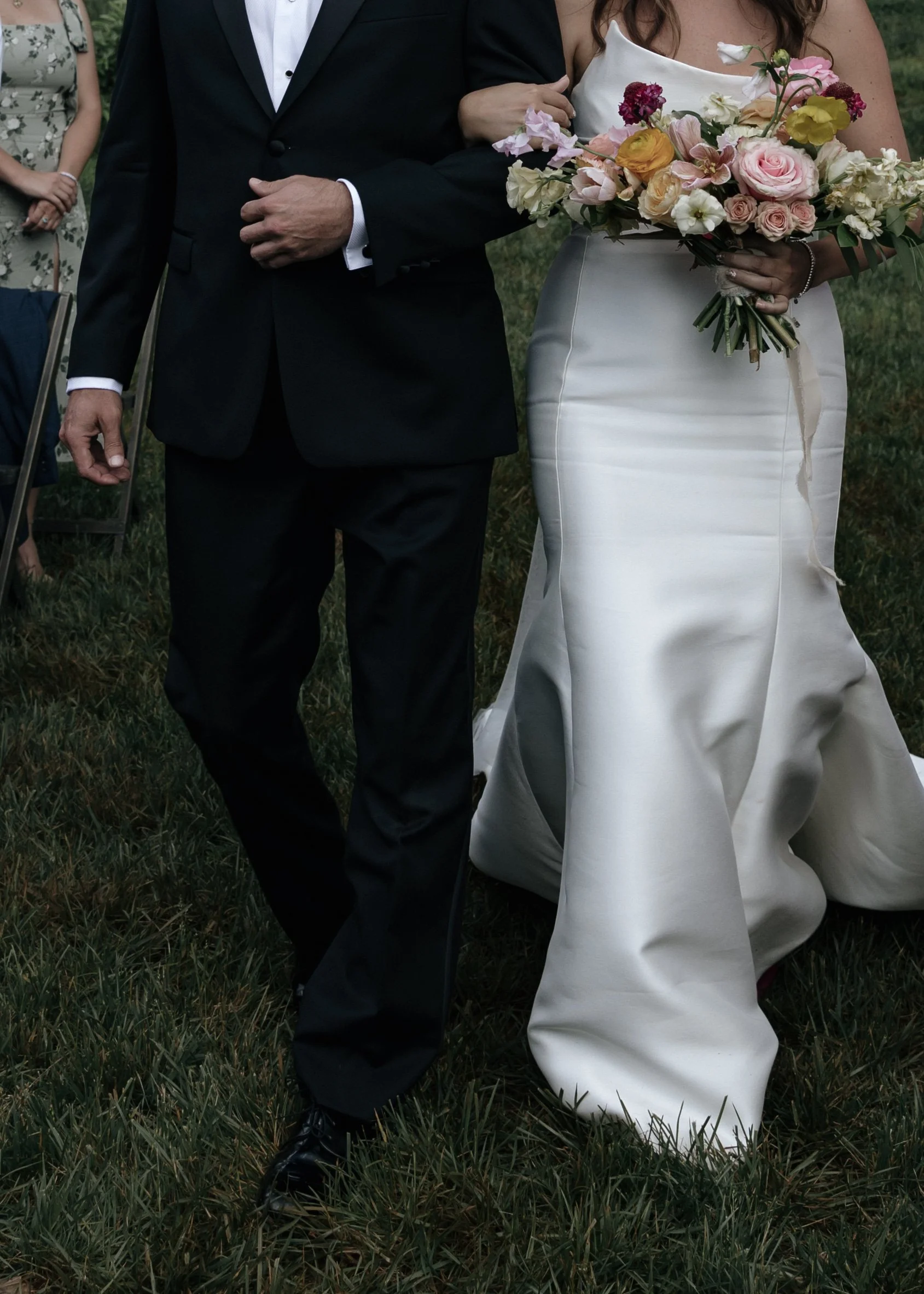 A bride in a white satin wedding dress holding a bouquet of pink, white, and yellow flowers, walking arm-in-arm with a man in a black suit during a wedding ceremony outdoors on grass.