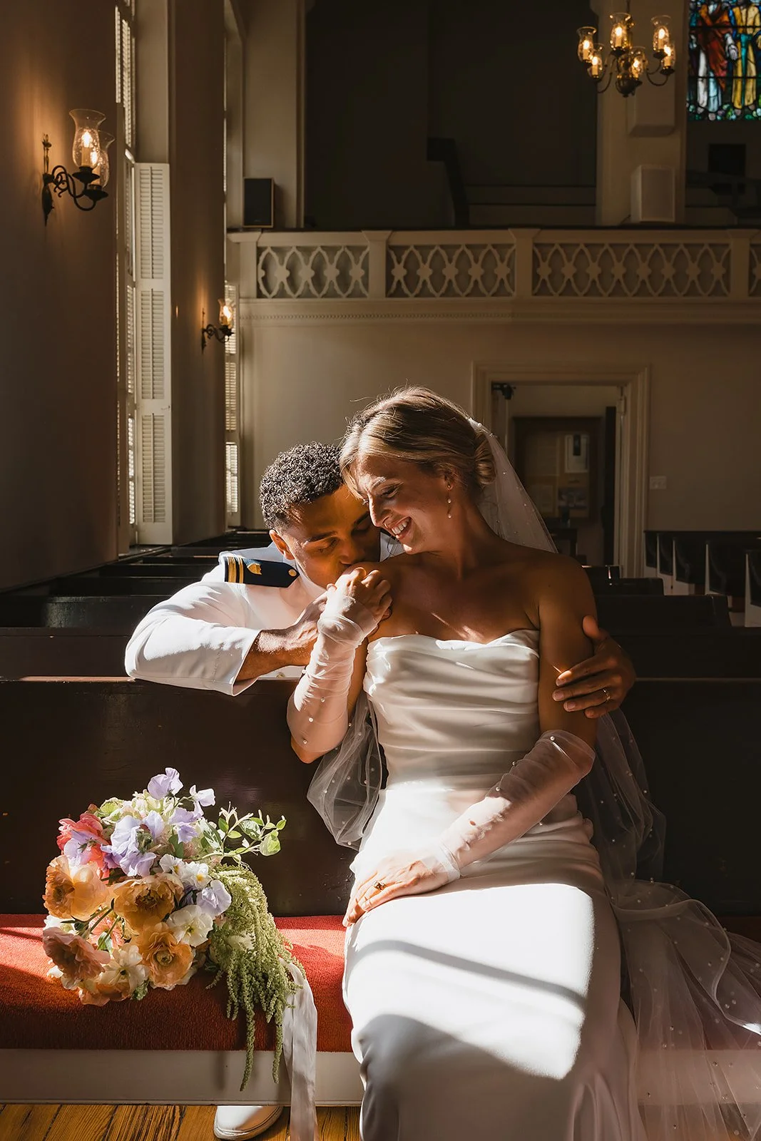 A bride and groom sitting in a church, sharing a joyful moment. The bride wears a white strapless wedding dress and veil, while the groom wears a white uniform with gold accents. They are smiling and leaning into each other, with the groom holding th