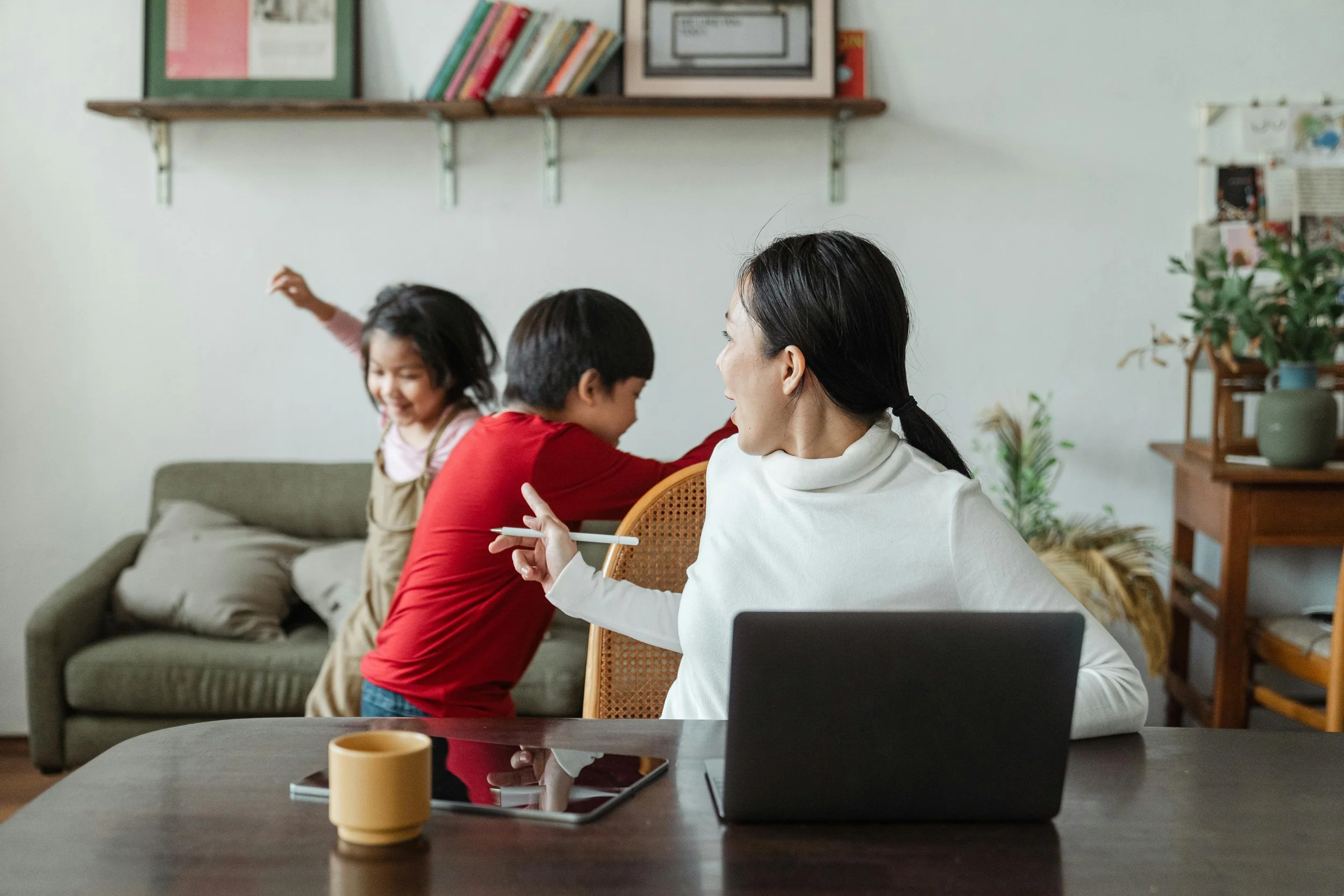 A woman is sitting at a table with a laptop and a tablet, pointing and talking to two children, a girl and a boy, who are standing behind her in a living room with a couch, plants, and shelves in the background.