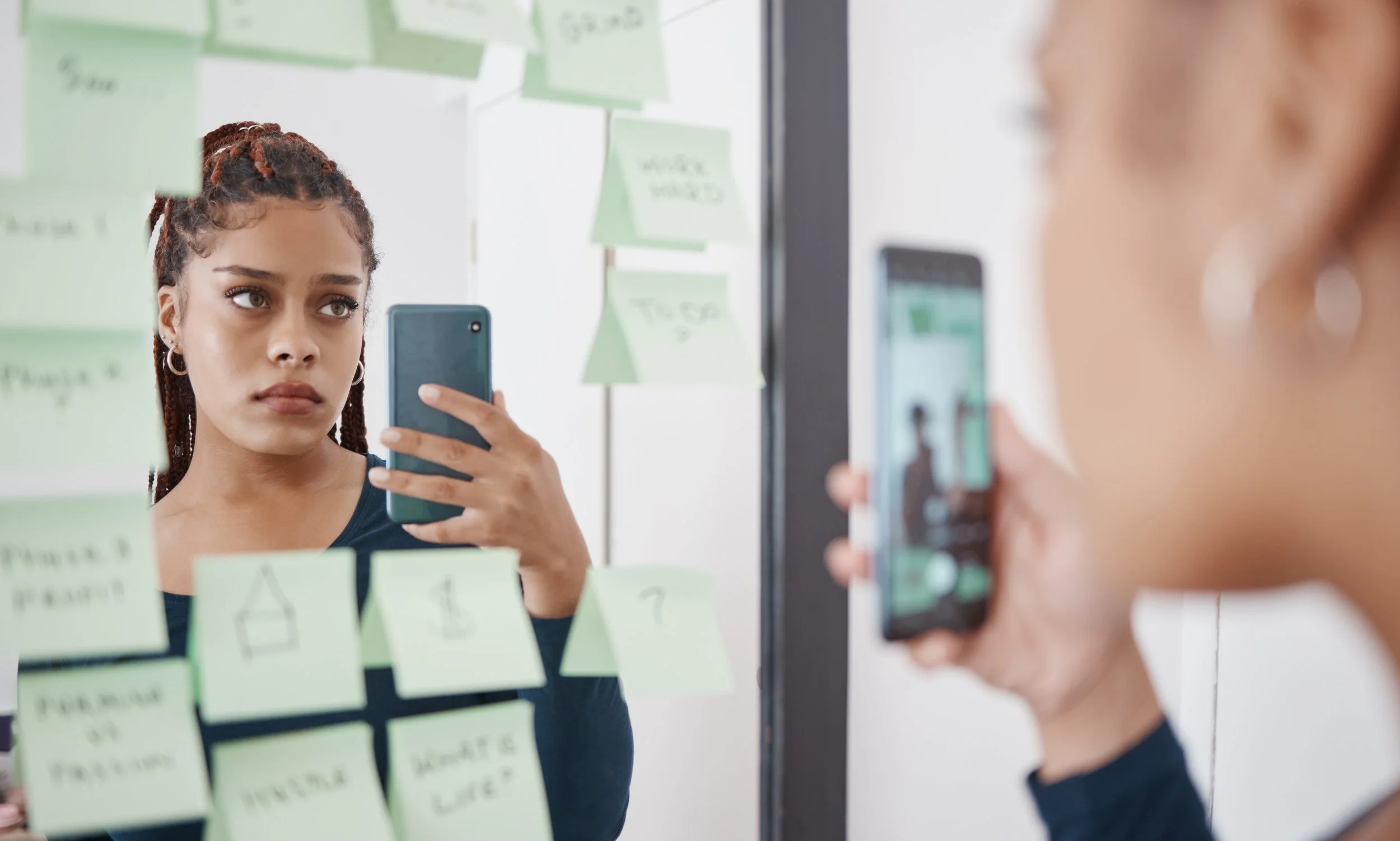 Young woman with braided hair and earrings taking a selfie in front of a mirror covered with green sticky notes with sketches and notes.