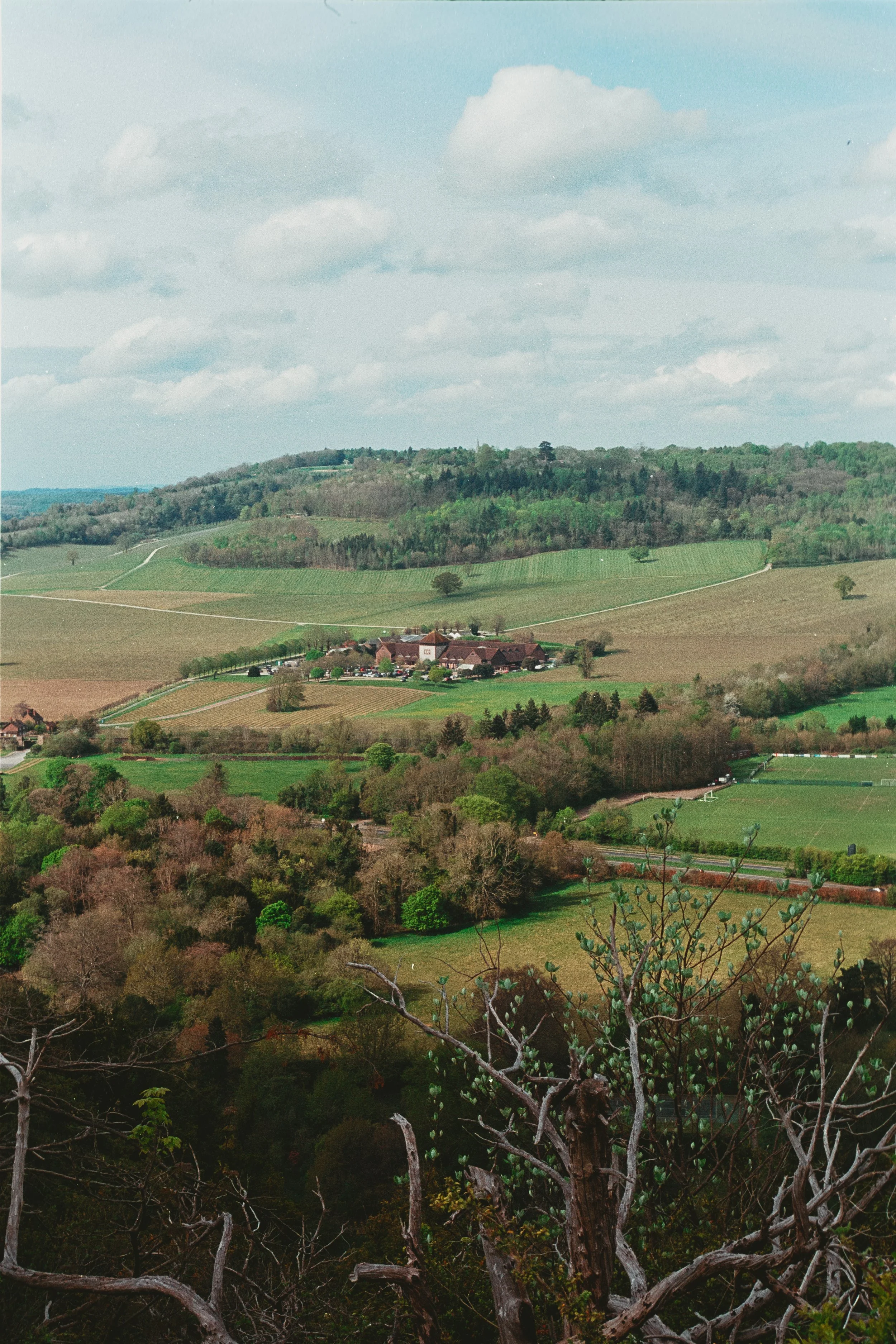 A scenic landscape of rolling hills and farmland with a small cluster of buildings, trees, and open fields under a partly cloudy sky.
