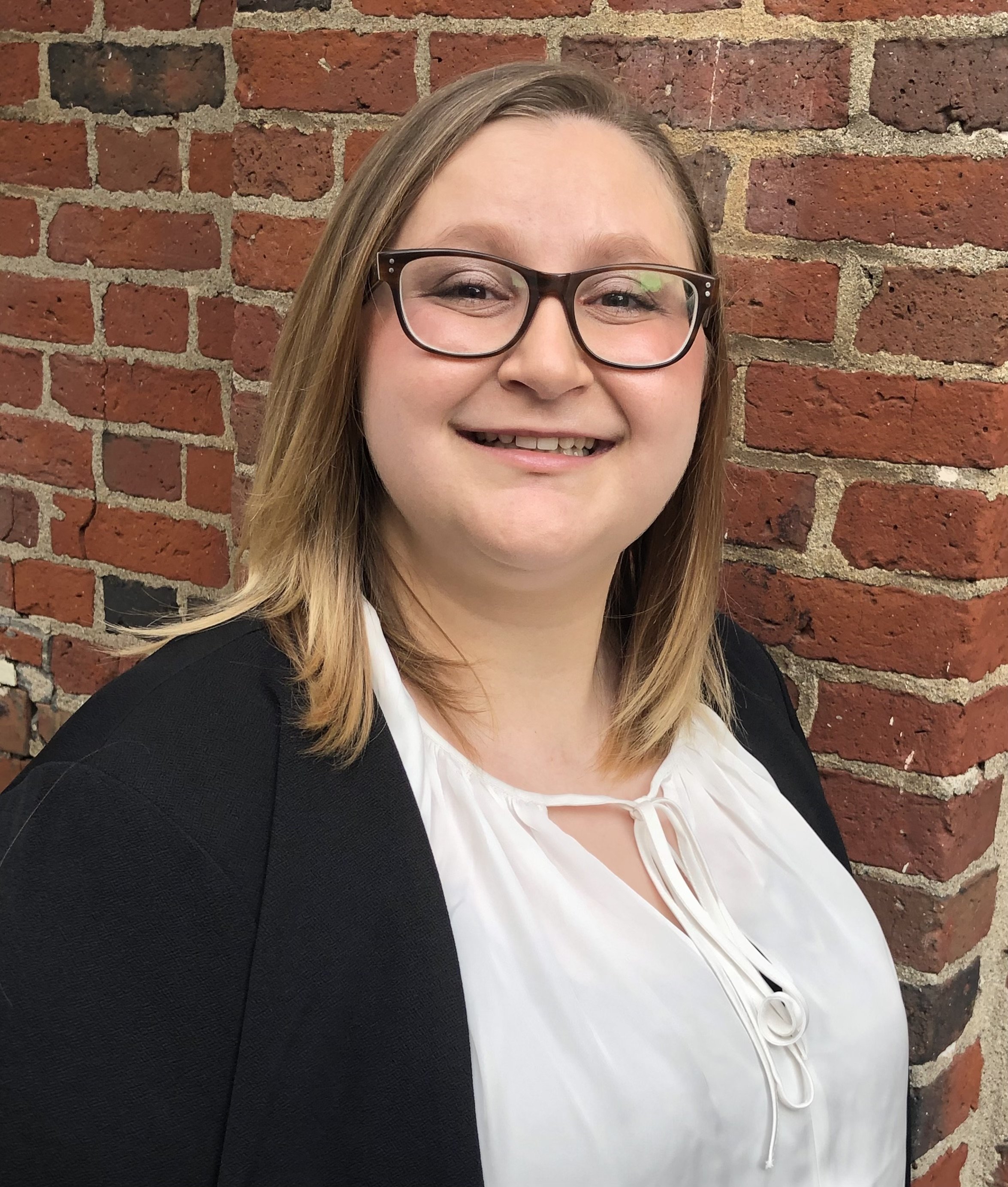 Retrato de una mujer con cabello largo castaño claro con gafas y una blusa naranja, de pie con los brazos cruzados contra un fondo oscuro.