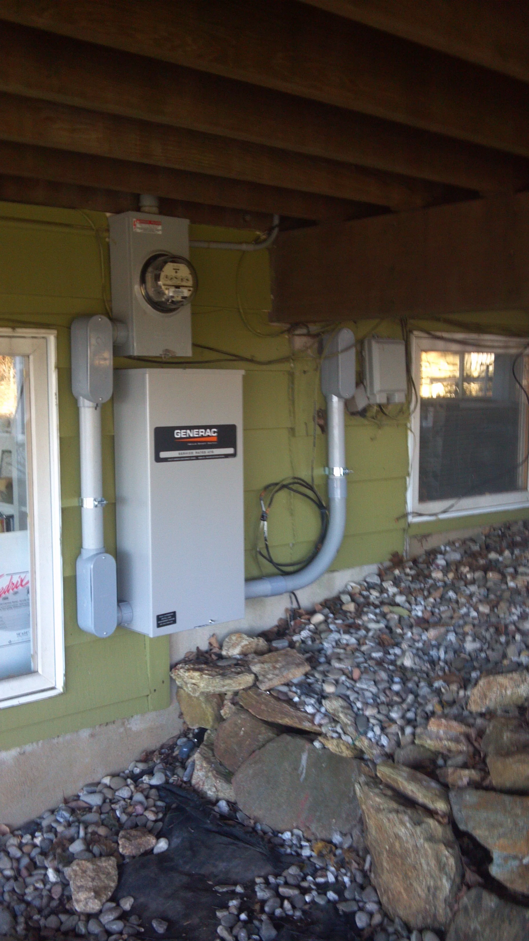 Exterior view of a house under a deck with a Generac generator, electrical meters, and wiring visible on the wall, surrounded by rocks and small stones.