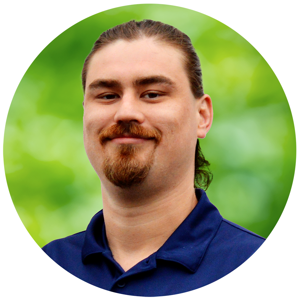 A man with long hair, mustache, and beard smiling outdoors with a green blurred background.
