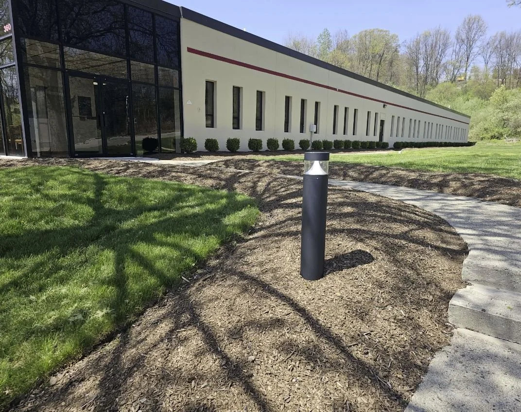 A modern building with large glass windows and a white exterior, situated on a well-maintained lawn with a curved concrete walkway and a black bollard light post in the foreground.