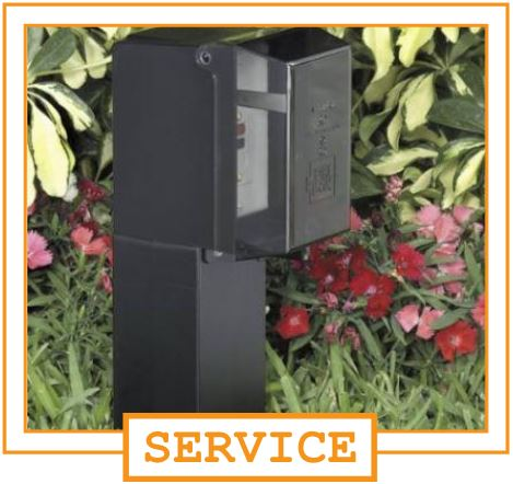 A black outdoor mailbox with a transparent front panel, mounted on a post, surrounded by pink and red flowers and green foliage.