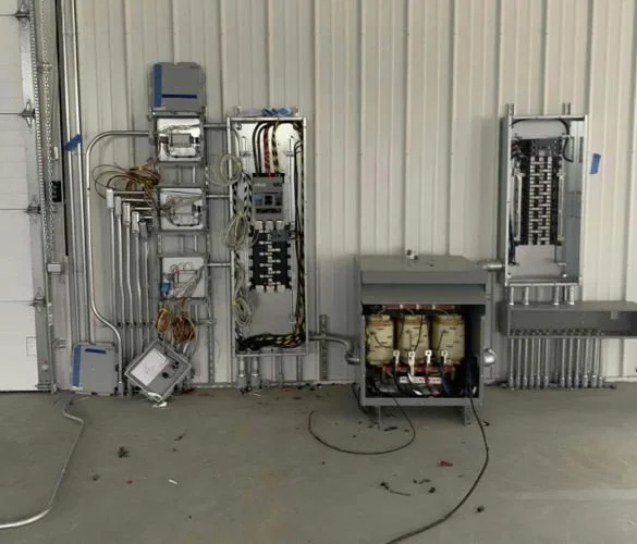 Electrical control panel with wiring and components mounted on a wall, including breakers, relays, and a transformer, in an industrial setting.