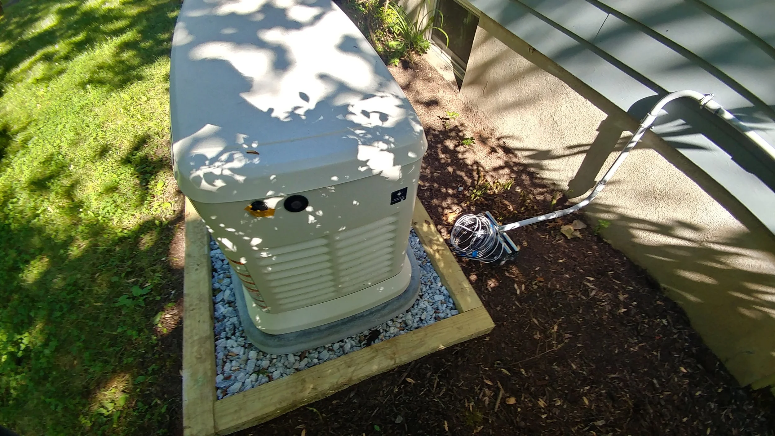 An outdoor air conditioning unit on a small gravel pad next to a house with a dirt strip along the foundation. There are shadows of tree branches on the unit and the ground.