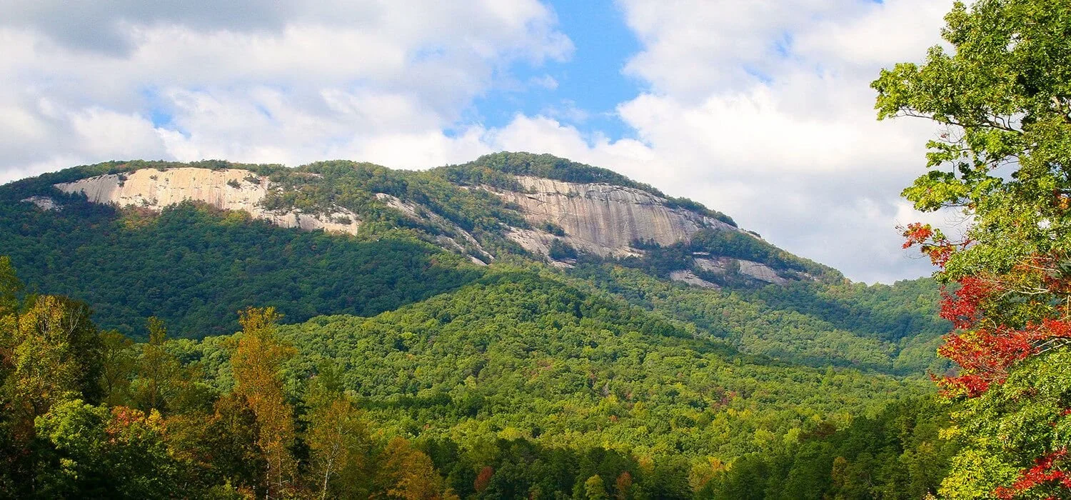 Sunlit mountain with layered rock formations and dense green forest in the foreground, cloudy sky above.