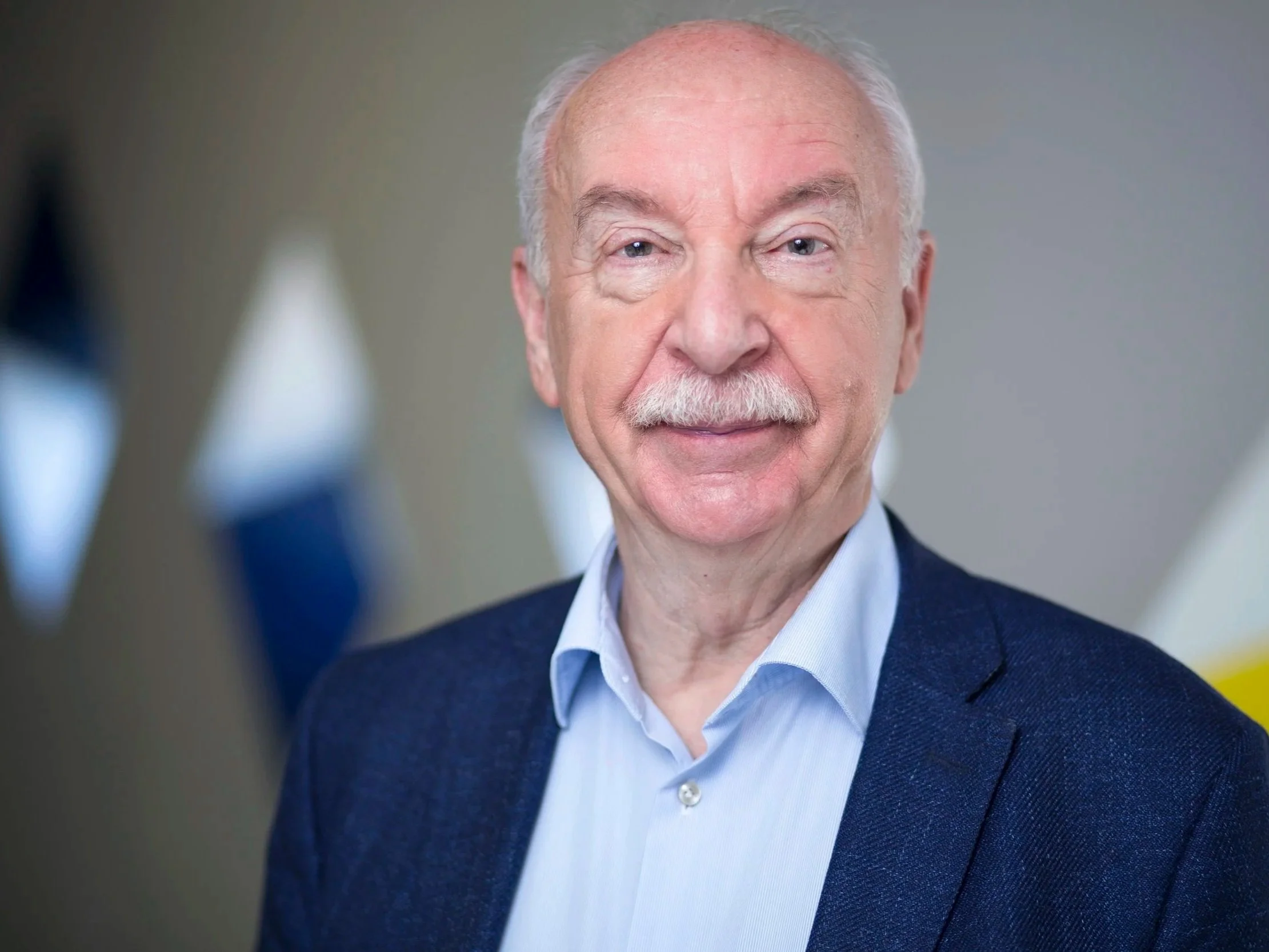 Close-up portrait of an elderly man with a white mustache, wearing a blue blazer and light blue dress shirt, smiling softly at the camera.