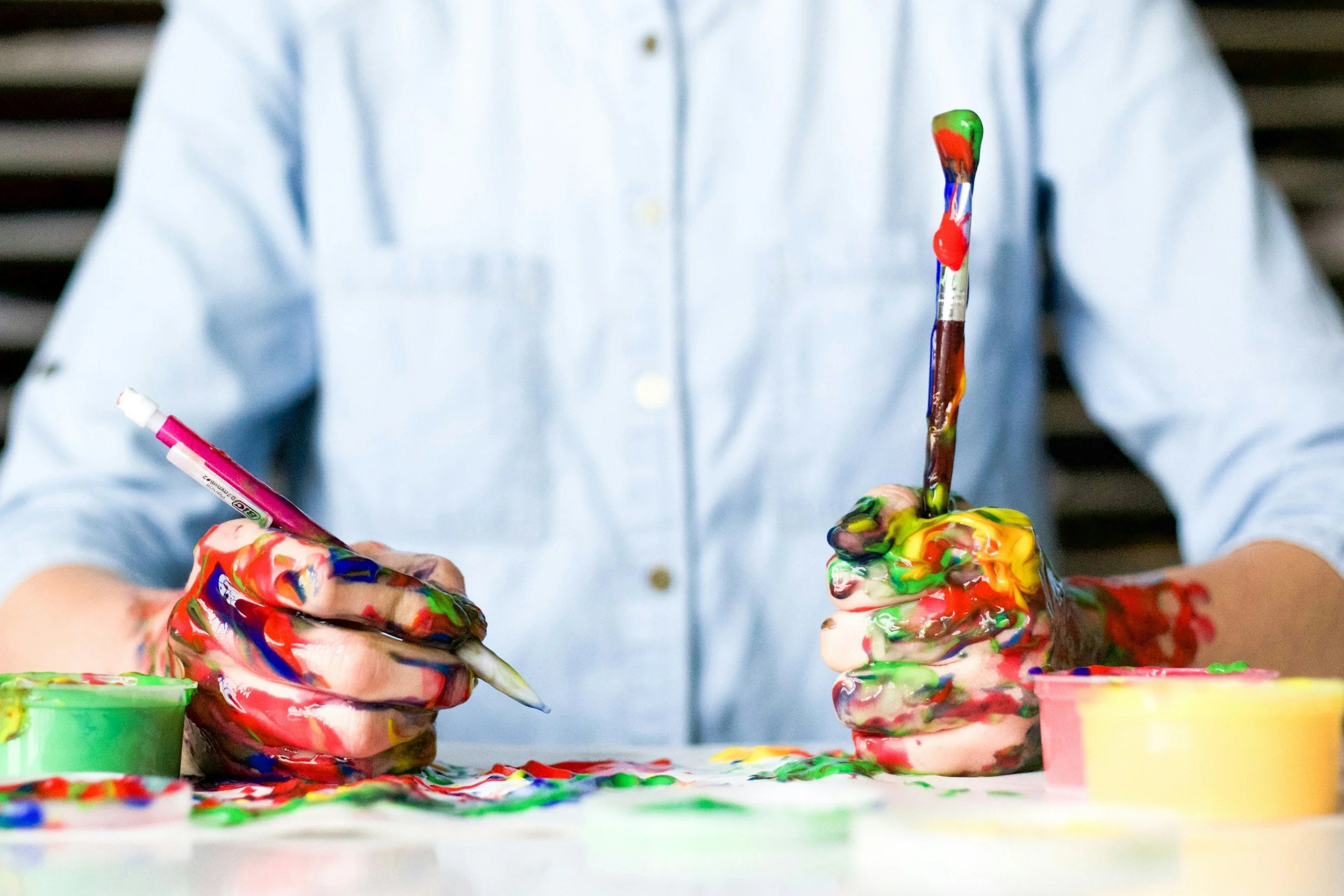 Person with hands covered in colorful paint holding a pen and a painted paintbrush, engaging in painting or artistic activity with paint containers in the foreground.