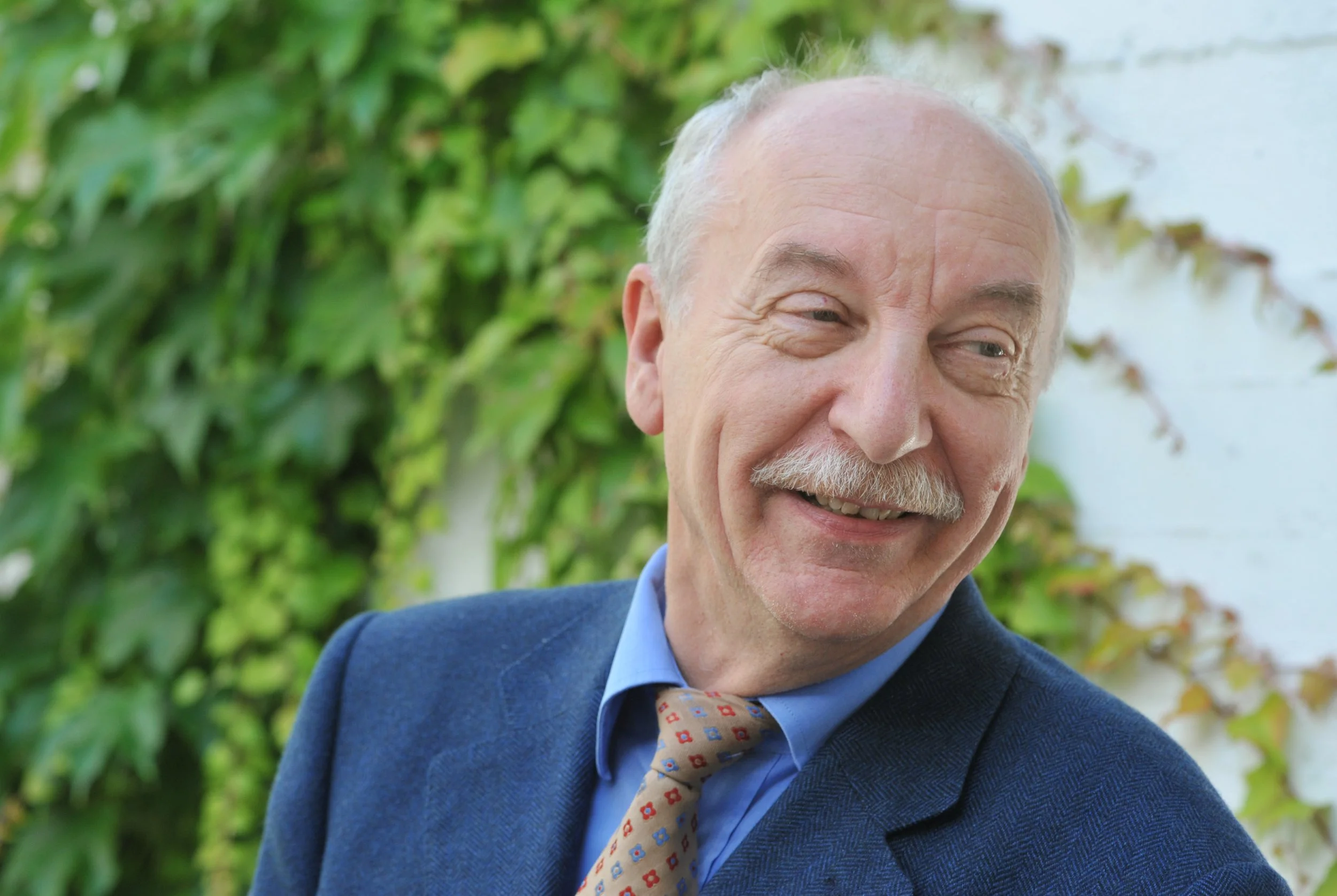 An elderly man smiling outdoors, wearing a blue suit and a patterned tie, with green foliage and a white wall in the background.