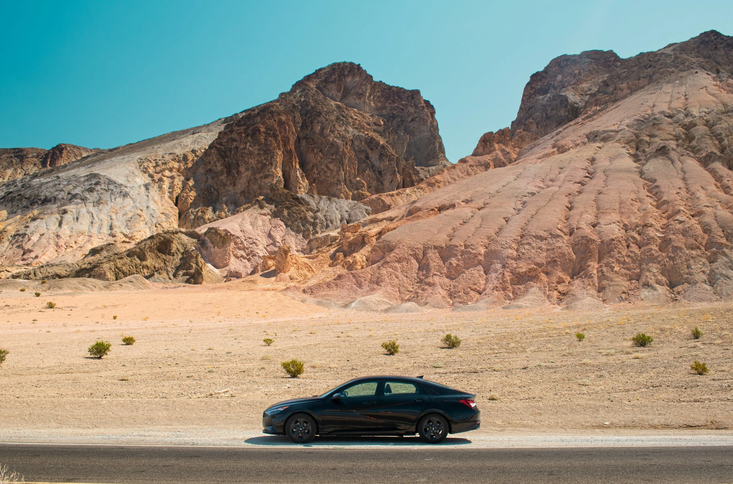 A black car parked on a roadside with a desert landscape and large rocky hills in the background under a clear blue sky.