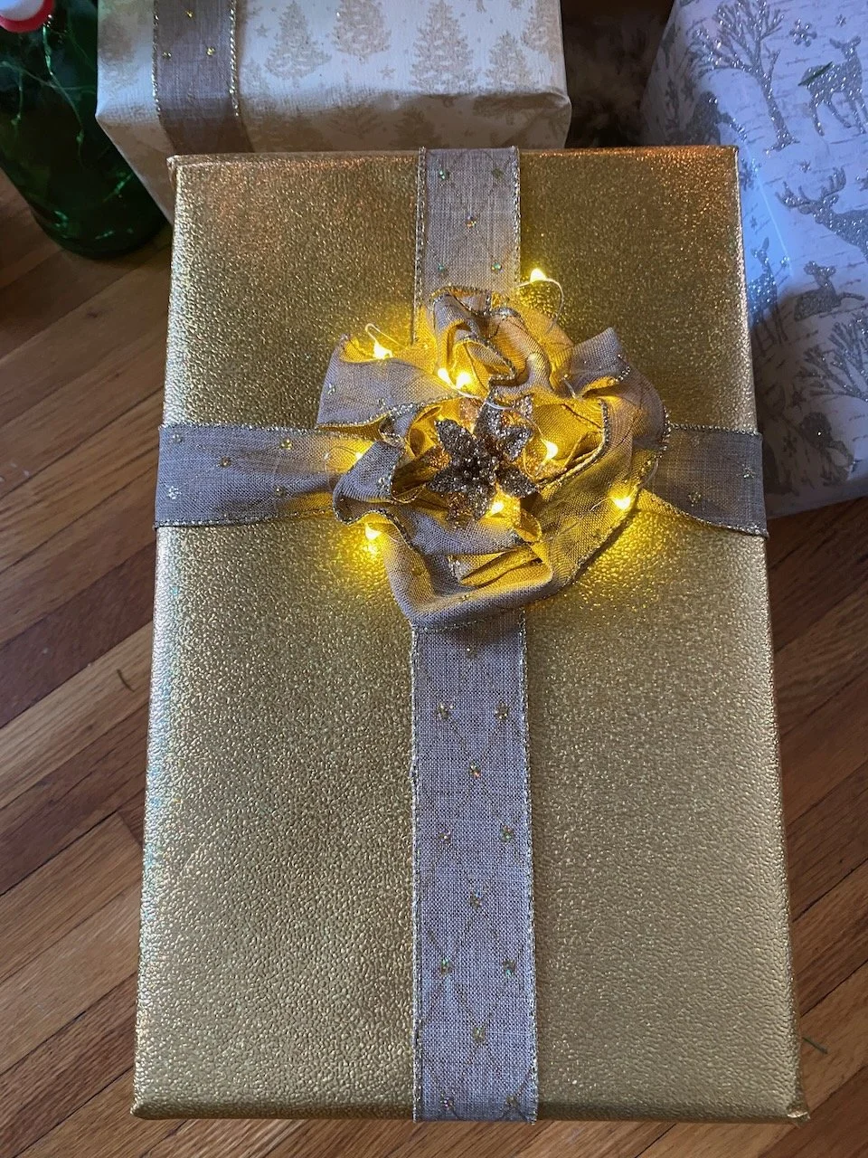 A Christmas gift wrapped in gold paper with a silver ribbon and decorated with a silver bow, illuminated by yellow string lights.