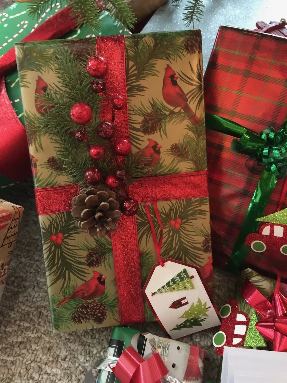 A Christmas gift box with holiday card design featuring cardinals, pine branches, pine cones, red berries, and red ribbon. Other wrapped presents are visible around it, decorated with festive bows and ribbons.