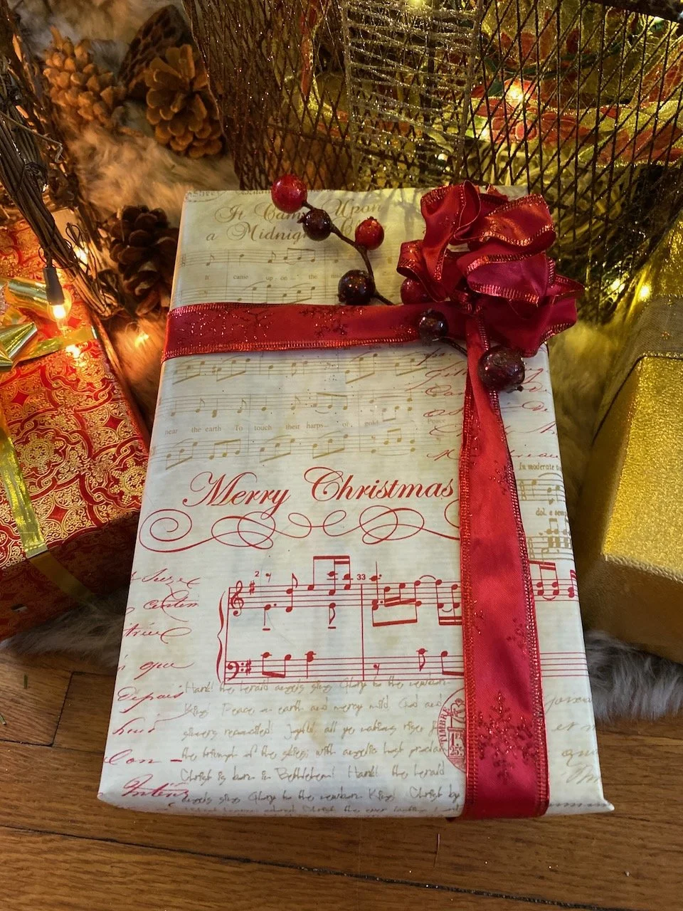 Decorative Christmas gift box wrapped in vintage sheet music paper design with a red ribbon and bow, surrounded by other wrapped presents, pinecones, and holiday decorations.