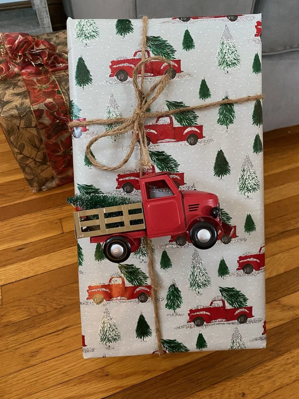 A Christmas gift wrapped in festive paper with red trucks and green trees, tied with a rustic twine bow, featuring a small red toy truck decoration on top.