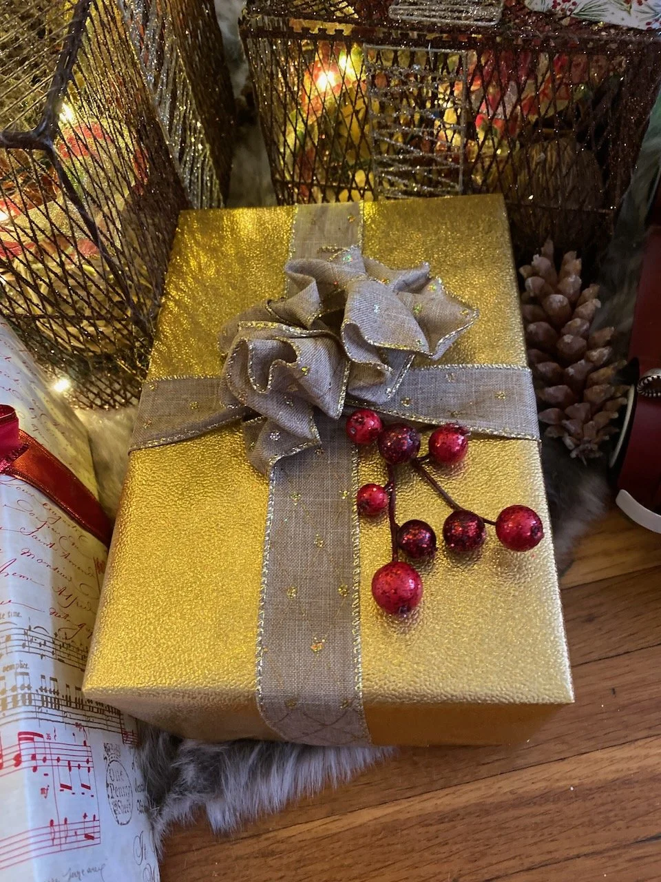 A brightly wrapped Christmas gift in gold and silver wrapping paper with a silver ribbon and bow, decorated with red berries, sitting on a wooden floor surrounded by other festive decorations.