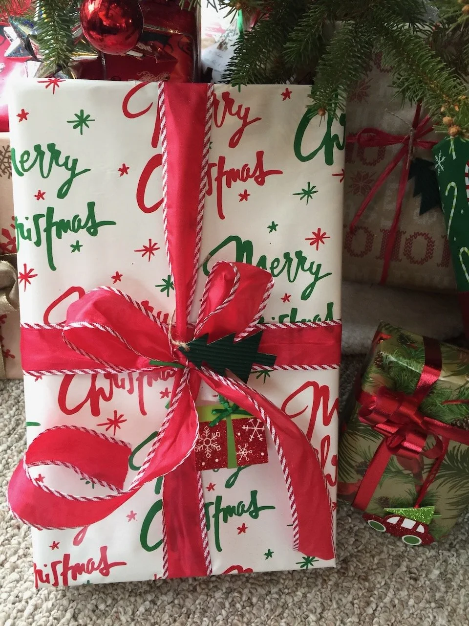Christmas gift wrapped in white paper with red and green 'Merry Christmas' text and small stars, decorated with a large red and white ribbon bow, placed under a Christmas tree with other wrapped presents nearby.