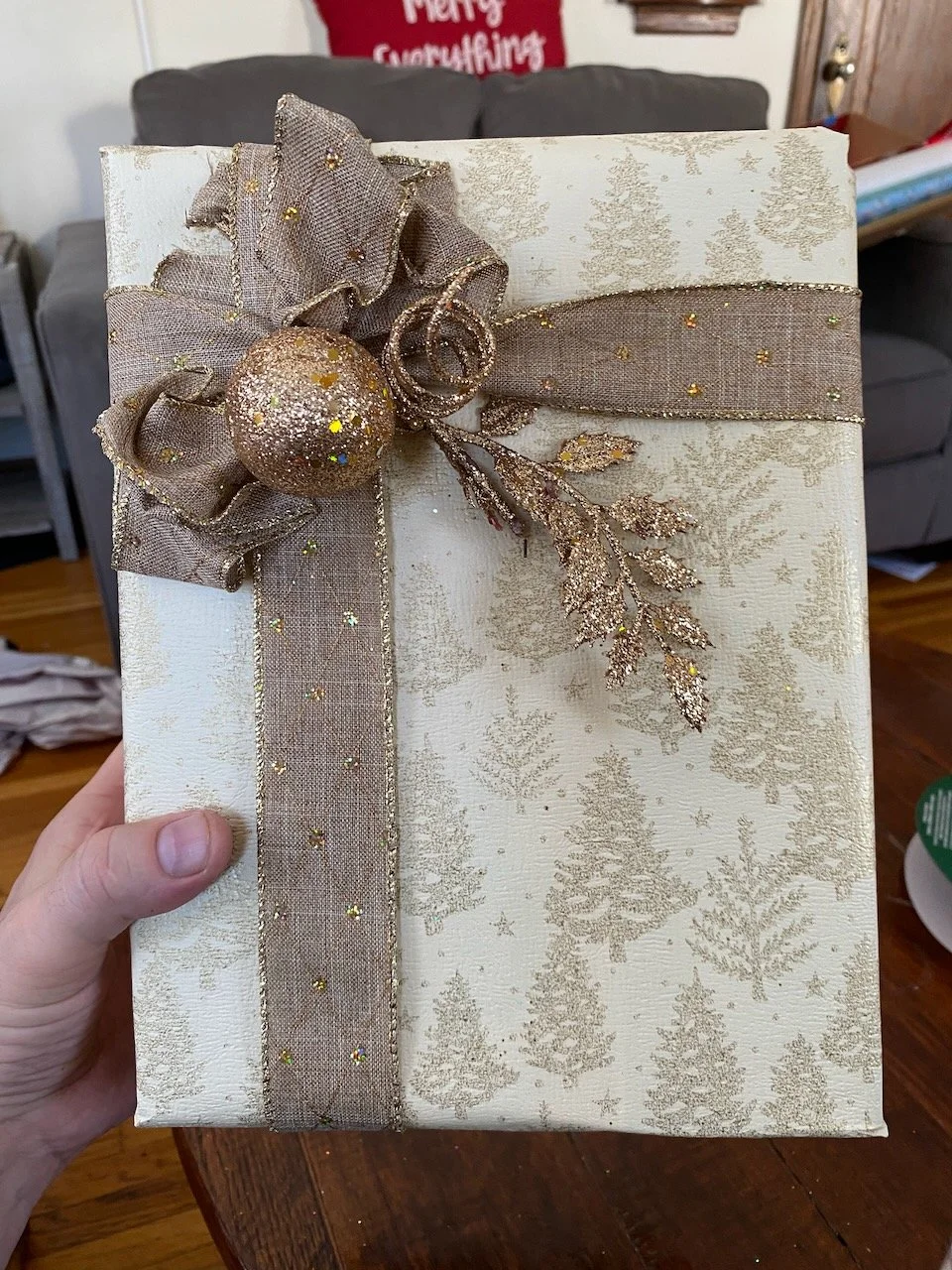 A Christmas gift wrapped in white and gold paper with a gold tree pattern, decorated with a gold ribbon, bow, a glittery gold ball, and glitter leaves.