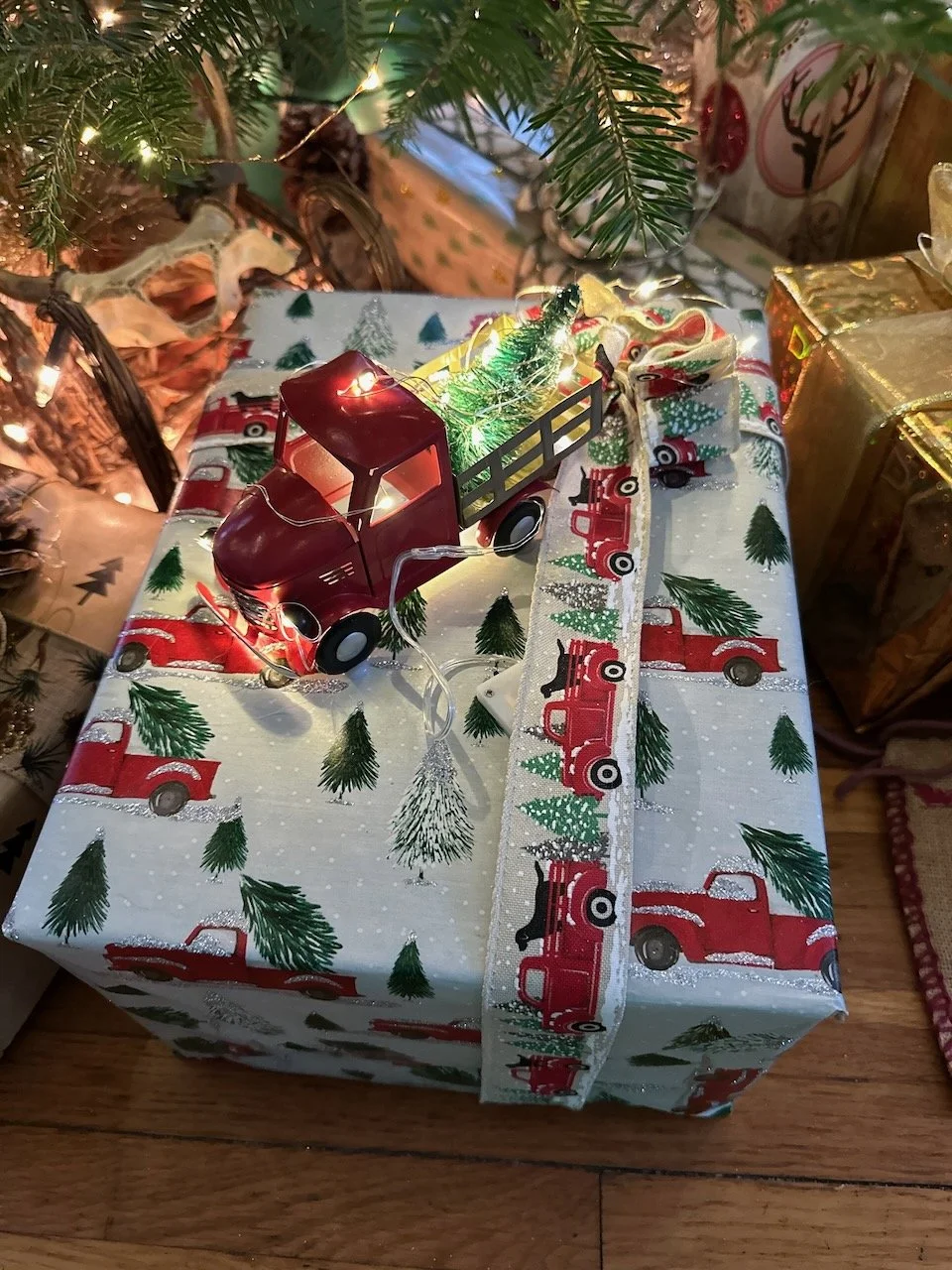 Festively wrapped Christmas gift with vintage red truck and Christmas tree wrapping paper, decorated with a small red toy truck, miniature Christmas tree, and a string of warm white fairy lights on top. Presents and Christmas tree visible nearby.