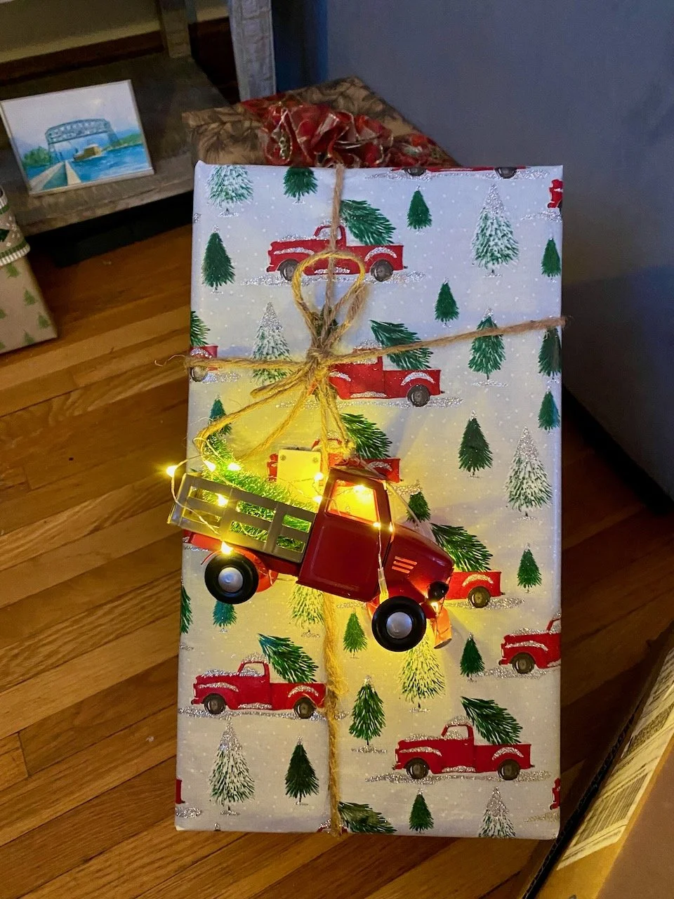 A Christmas gift wrapped in festive paper with a pattern of red trucks and green trees, decorated with a small red toy truck, yellow string lights, and tied with a yellow string.